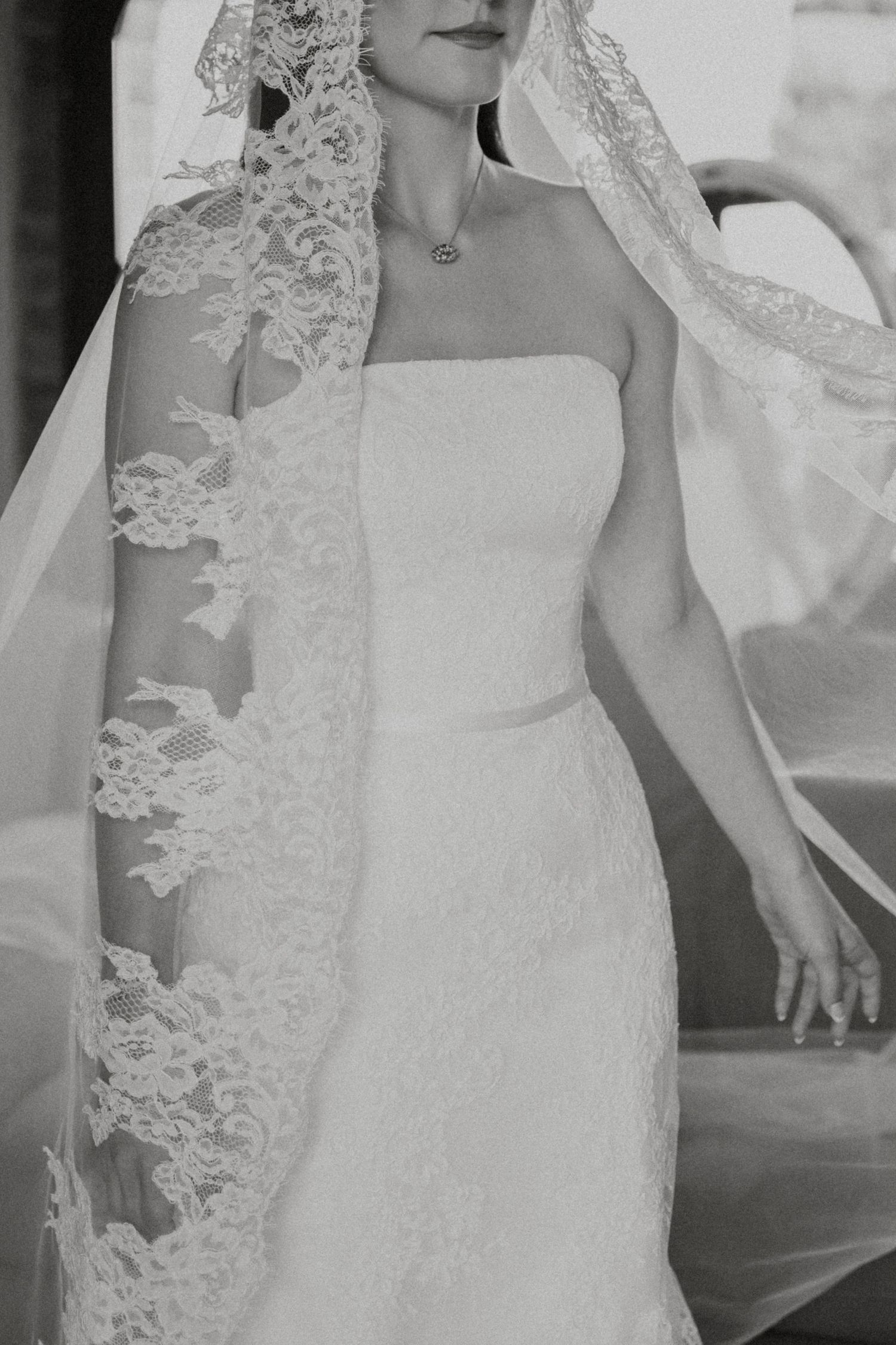 Bride in strapless lace wedding gown wears ornate lace mantilla veil in black and white portrait.
