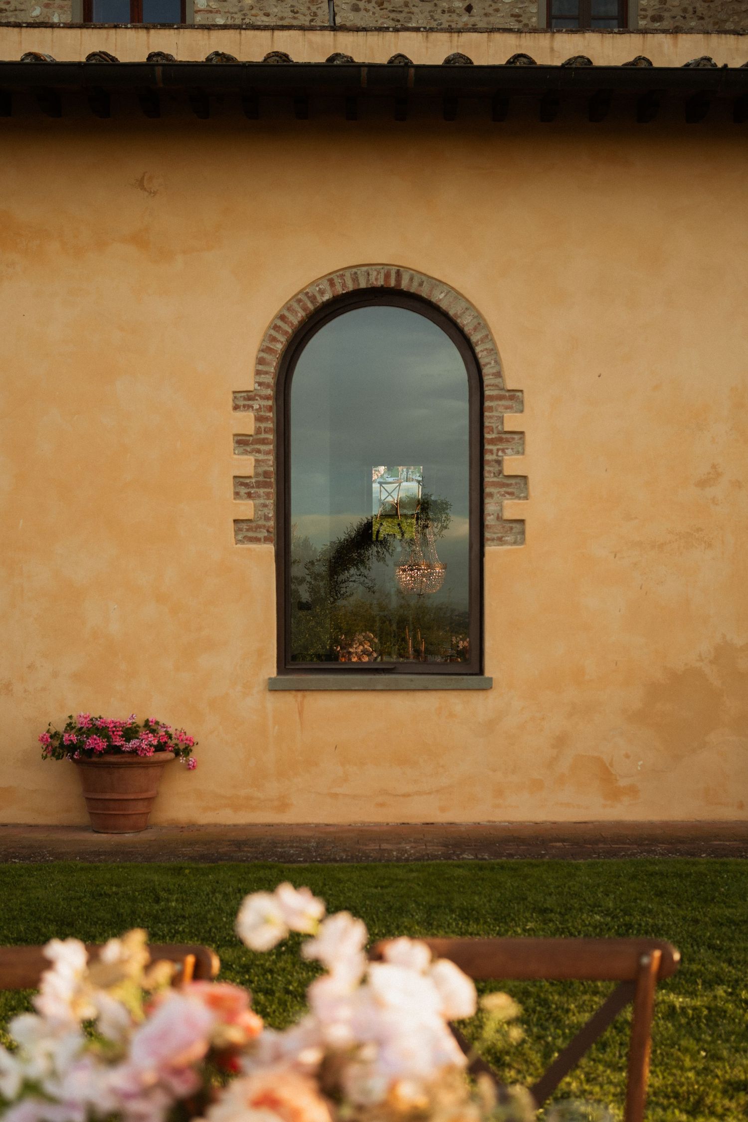 Arched window on rustic Tuscan villa wall with terracotta pots and soft floral arrangements in foreground.