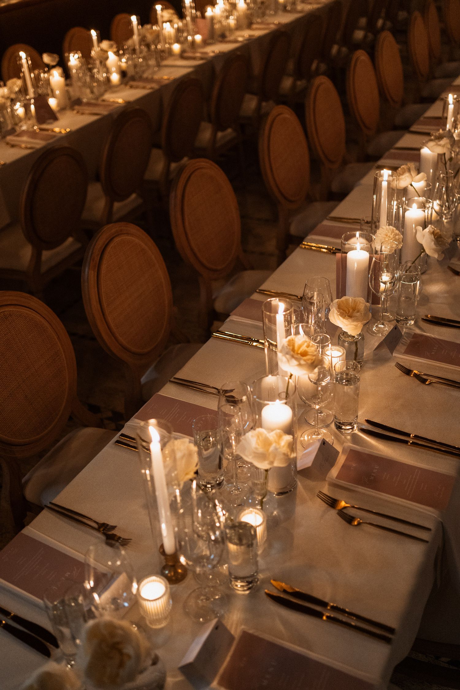 Elegant wedding reception table set with candles, white roses, crystal glassware, and gold cutlery.