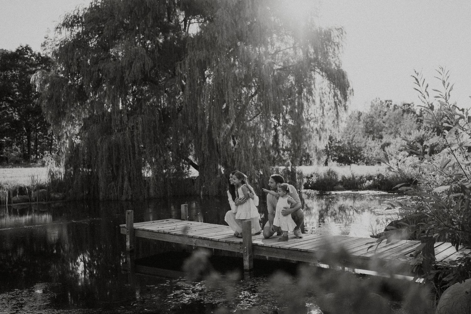 Black and white photo of a family sitting together on a wooden dock beside a tranquil pond under a weeping willow.