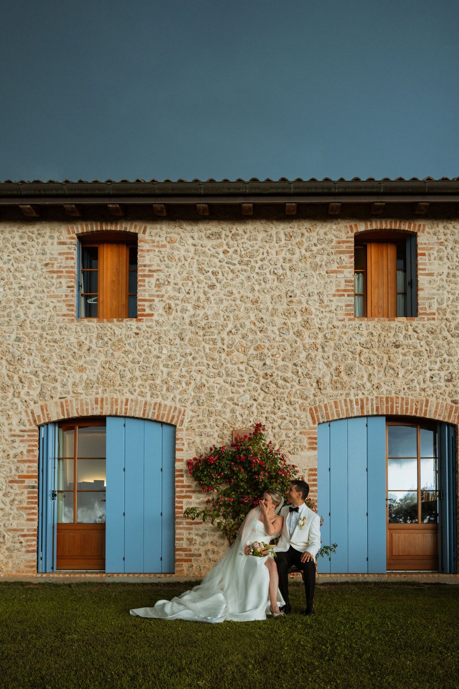 Bride and groom kissing in front of a rustic stone building with blue shutters and climbing roses.