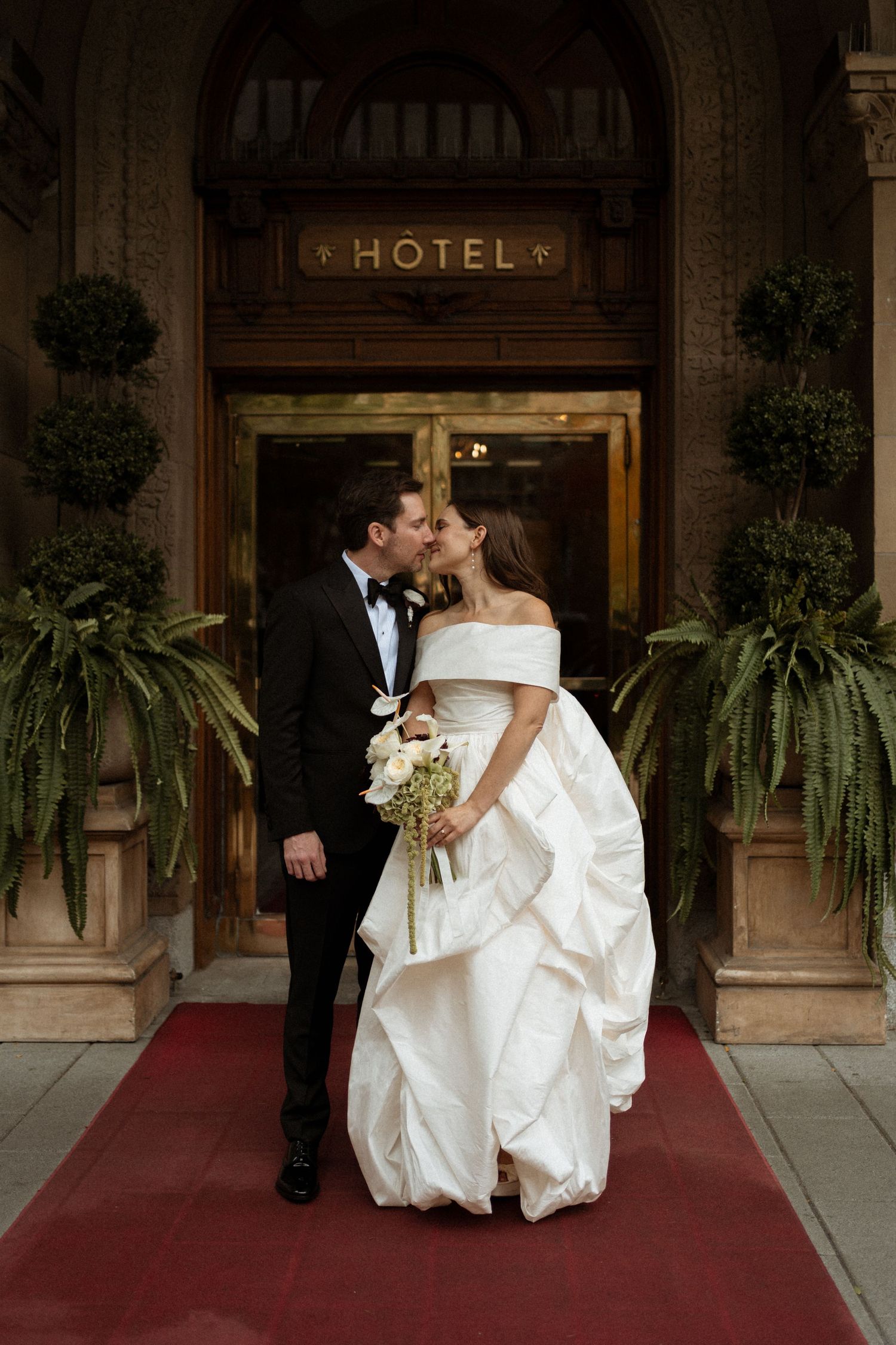 Bride and groom kiss on red carpet outside elegant hotel entrance, bride in voluminous off-shoulder white gown.