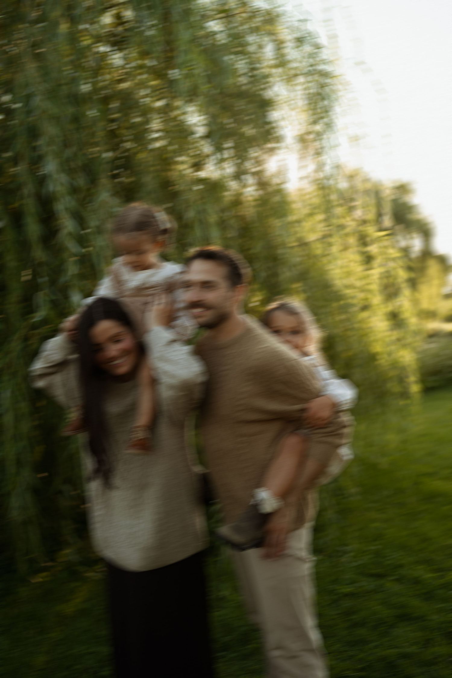 A happy family with two young children enjoys a sunny day outdoors near lush green weeping willow trees.