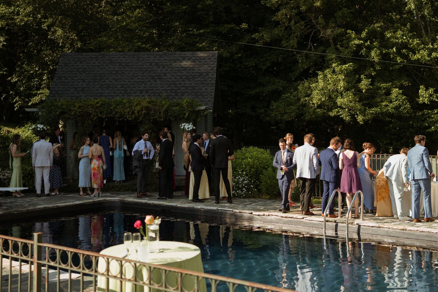 Elegant outdoor wedding reception with guests mingling beside a reflective pool surrounded by lush green trees.