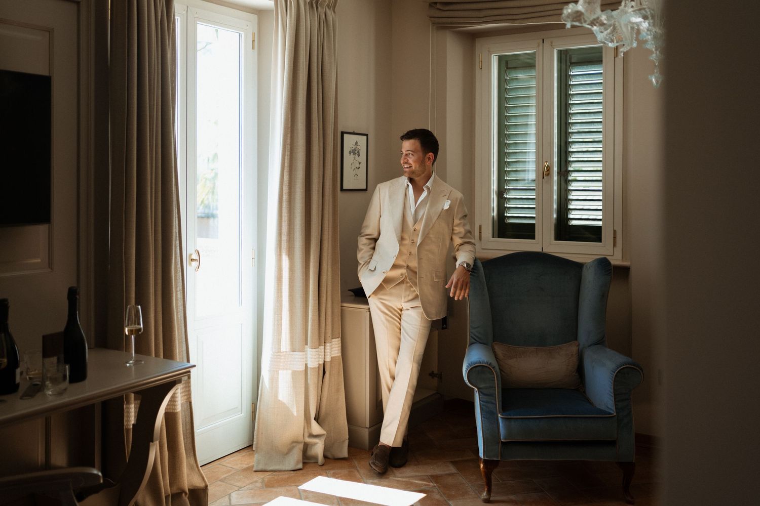 Elegant man in cream suit standing by sunlit French doors in a luxurious room with blue armchair.