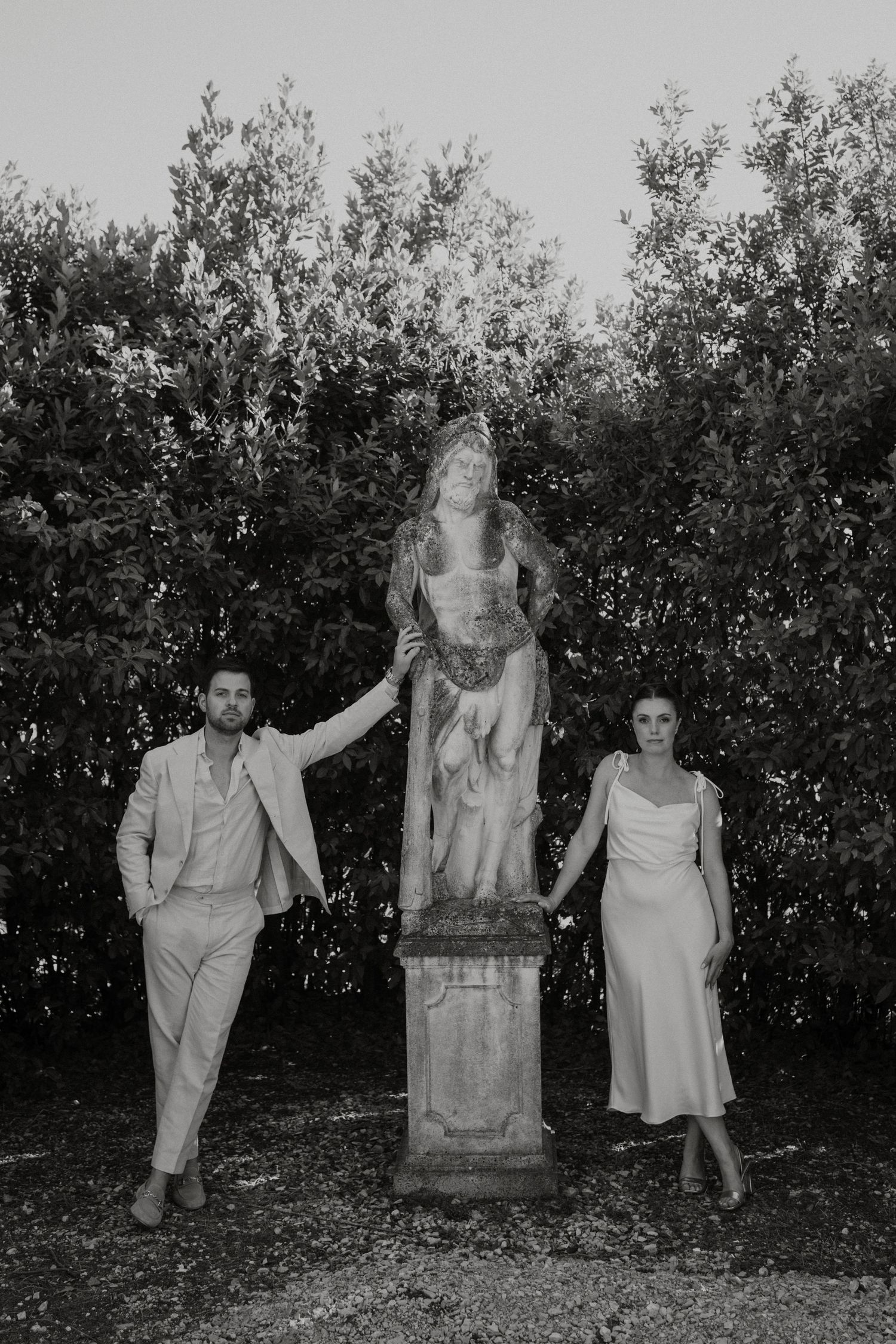 Black and white photo of a couple posing beside an ornate garden statue surrounded by lush trees.