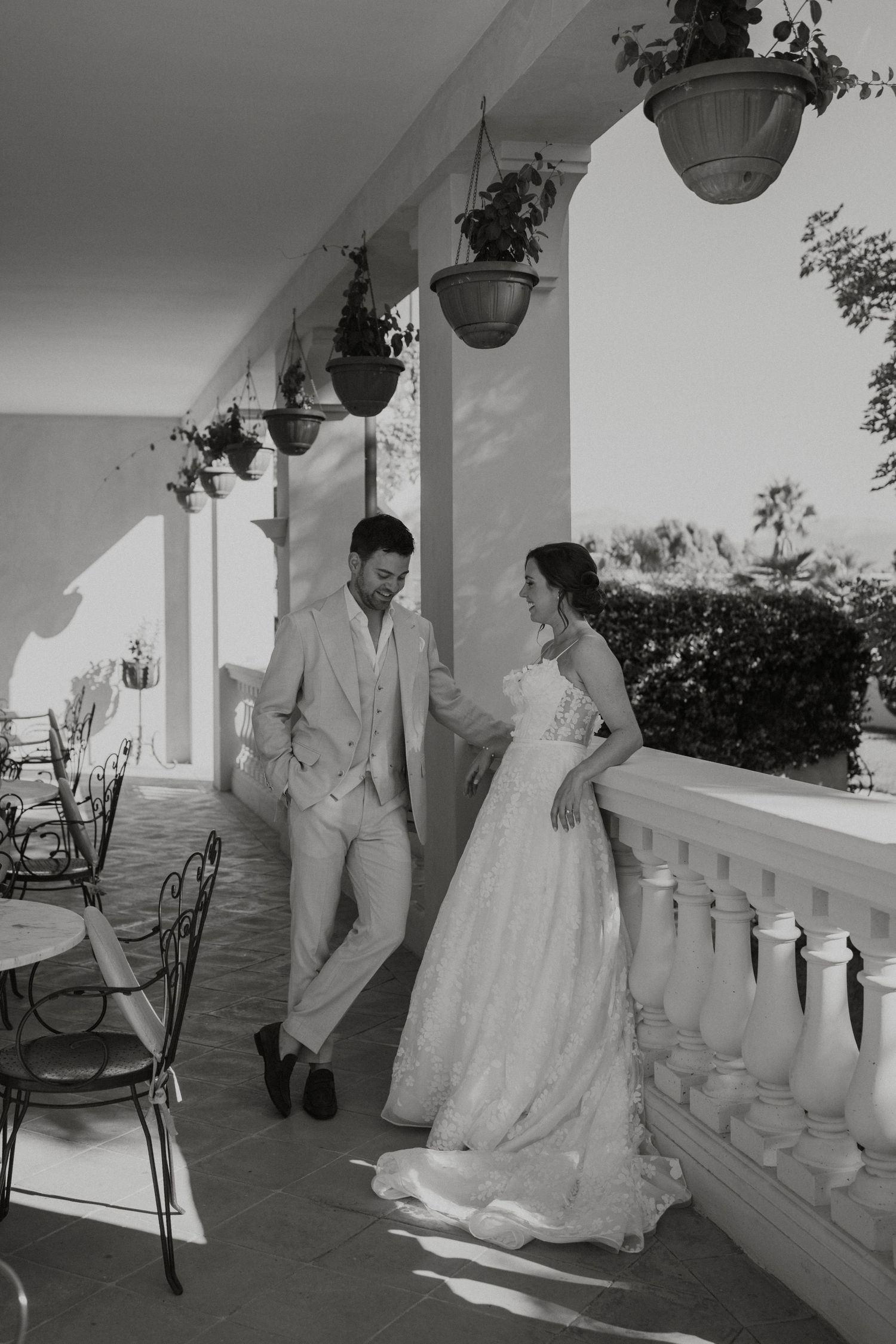 Black and white photo of bride and groom on sunlit villa terrace with hanging plants and ornate balustrade.