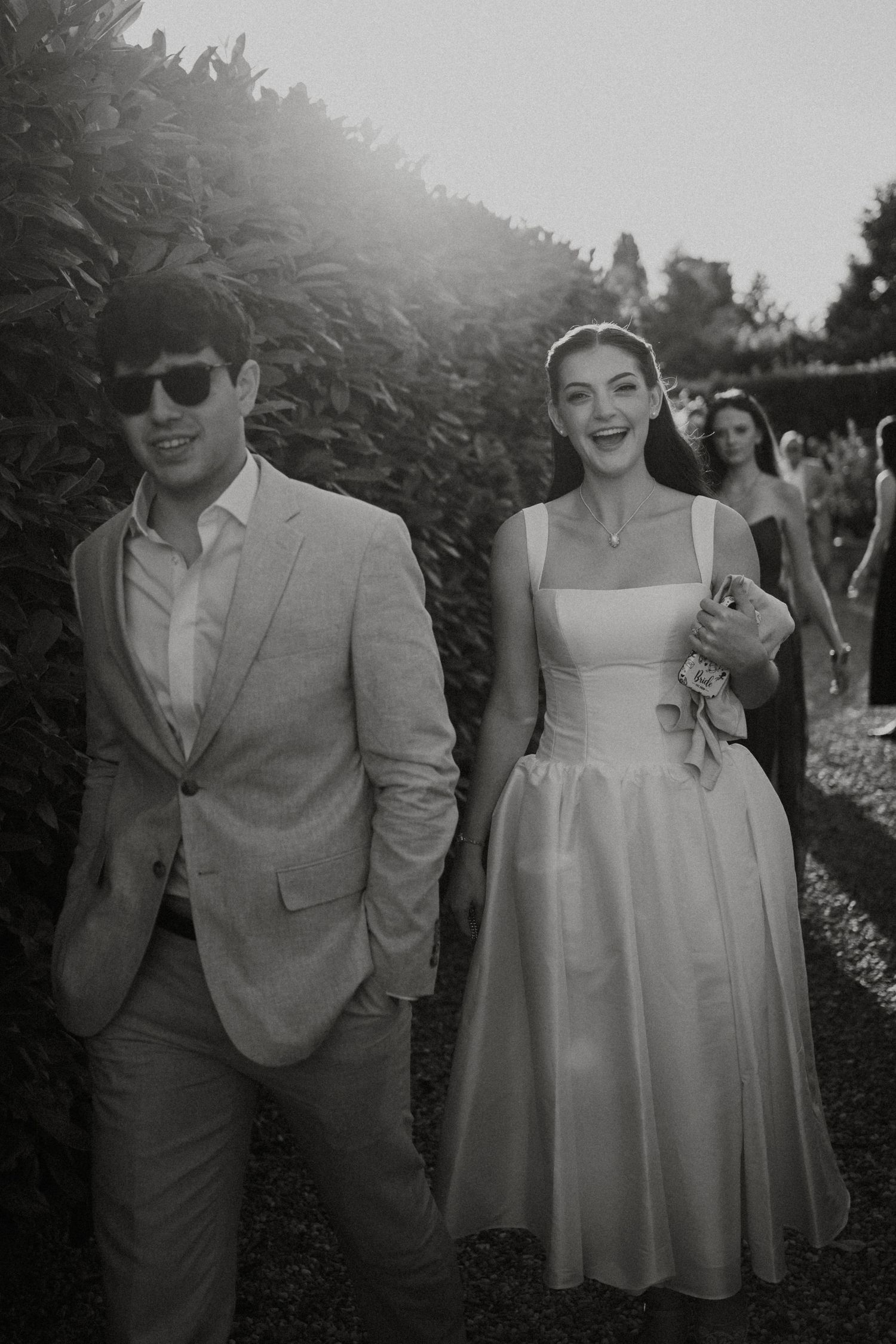 Smiling couple walks through garden, man in grey suit with sunglasses, woman in white square-neck midi dress.