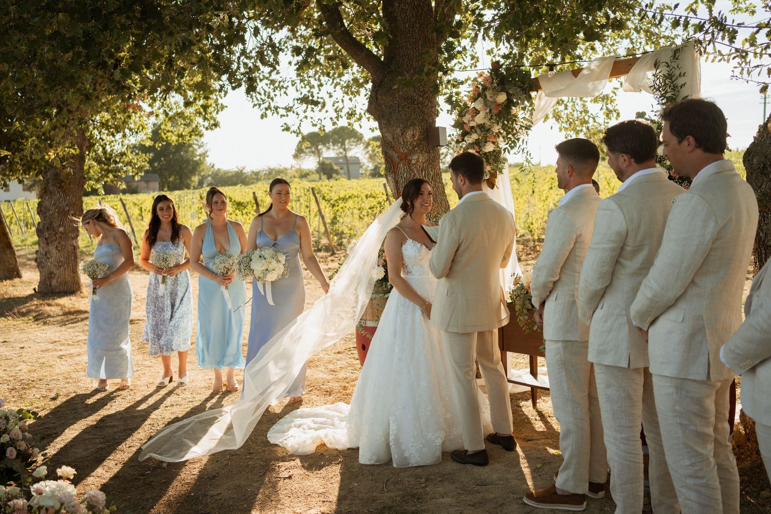 Outdoor vineyard wedding ceremony with bride in white gown, bridesmaids in blue dresses, and groomsmen in cream suits.