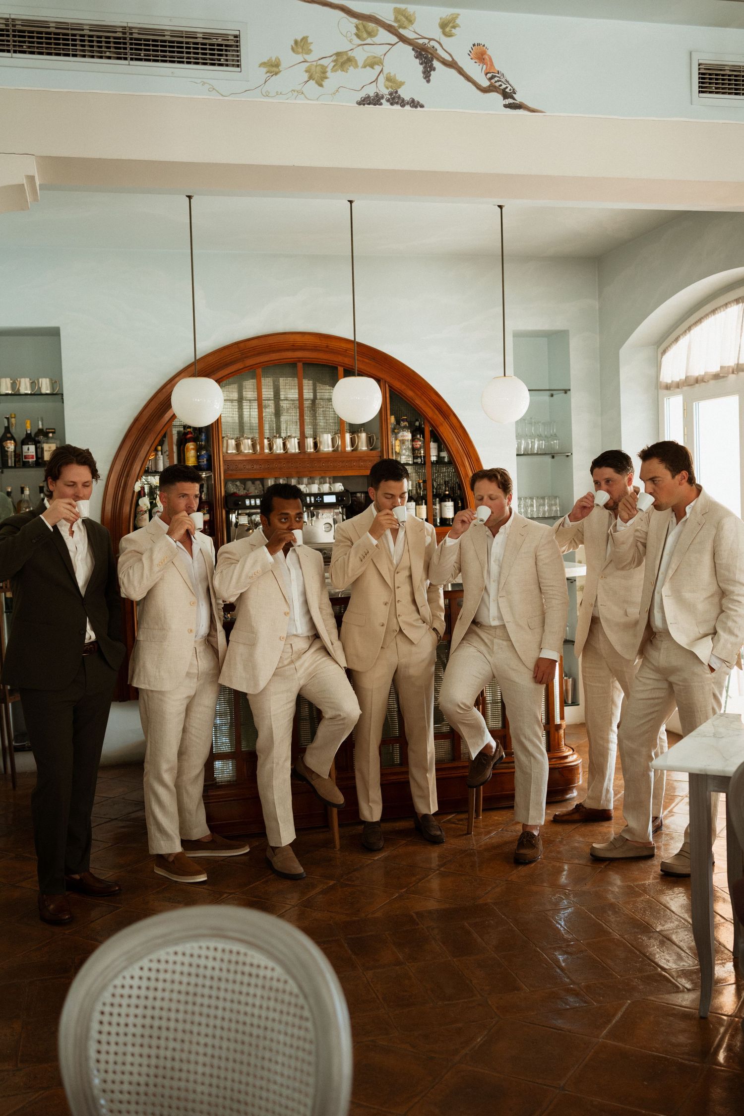 Groomsmen in matching beige suits laughing and toasting drinks at an elegant bar venue.