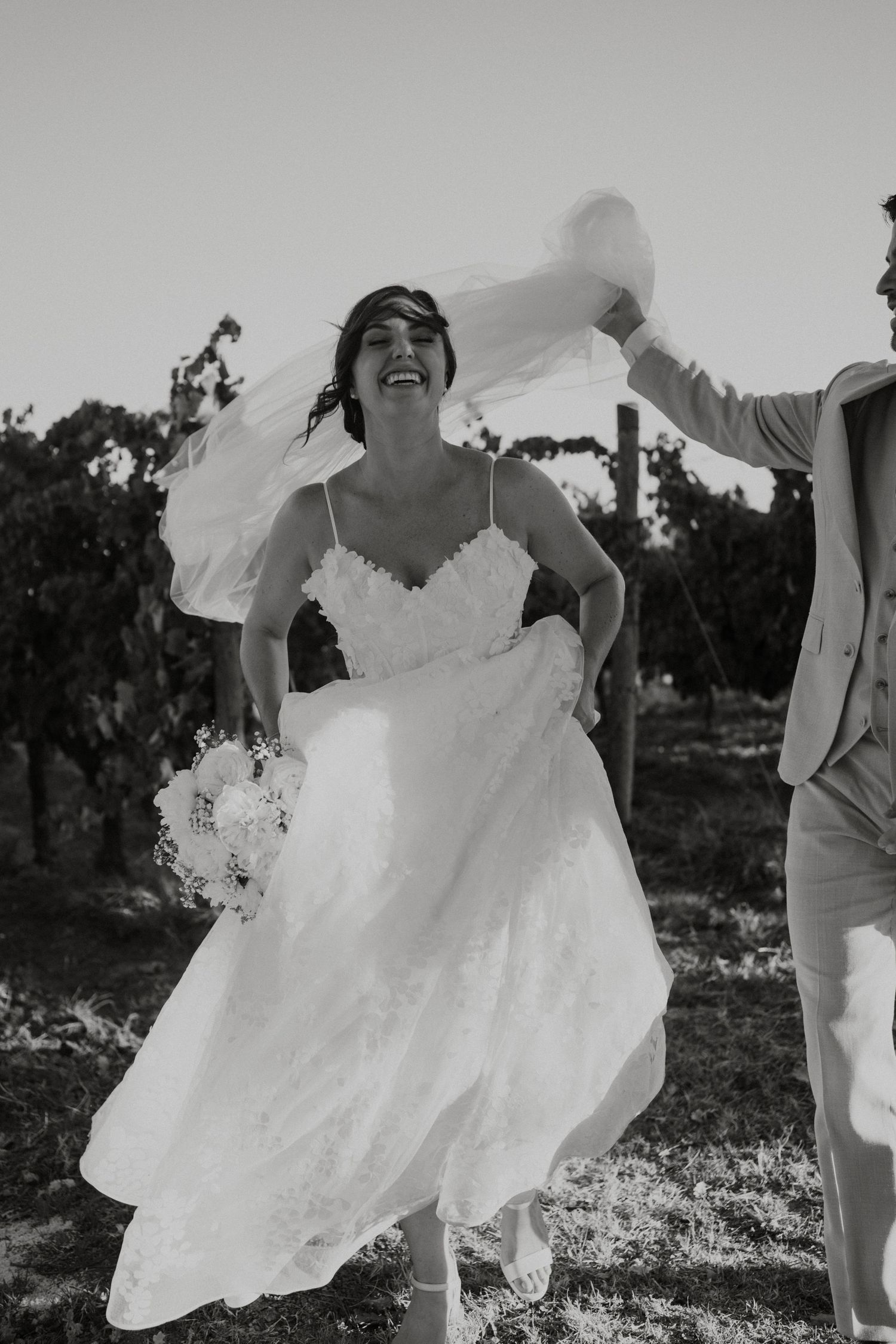 Black and white photo of laughing bride in lace gown holding bouquet while veil flows in the wind at vineyard.