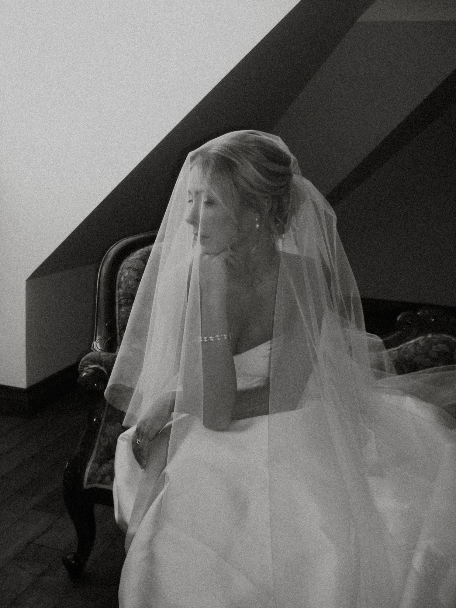 Elegant black and white portrait of bride in wedding gown and veil seated in vintage chair near slanted ceiling.