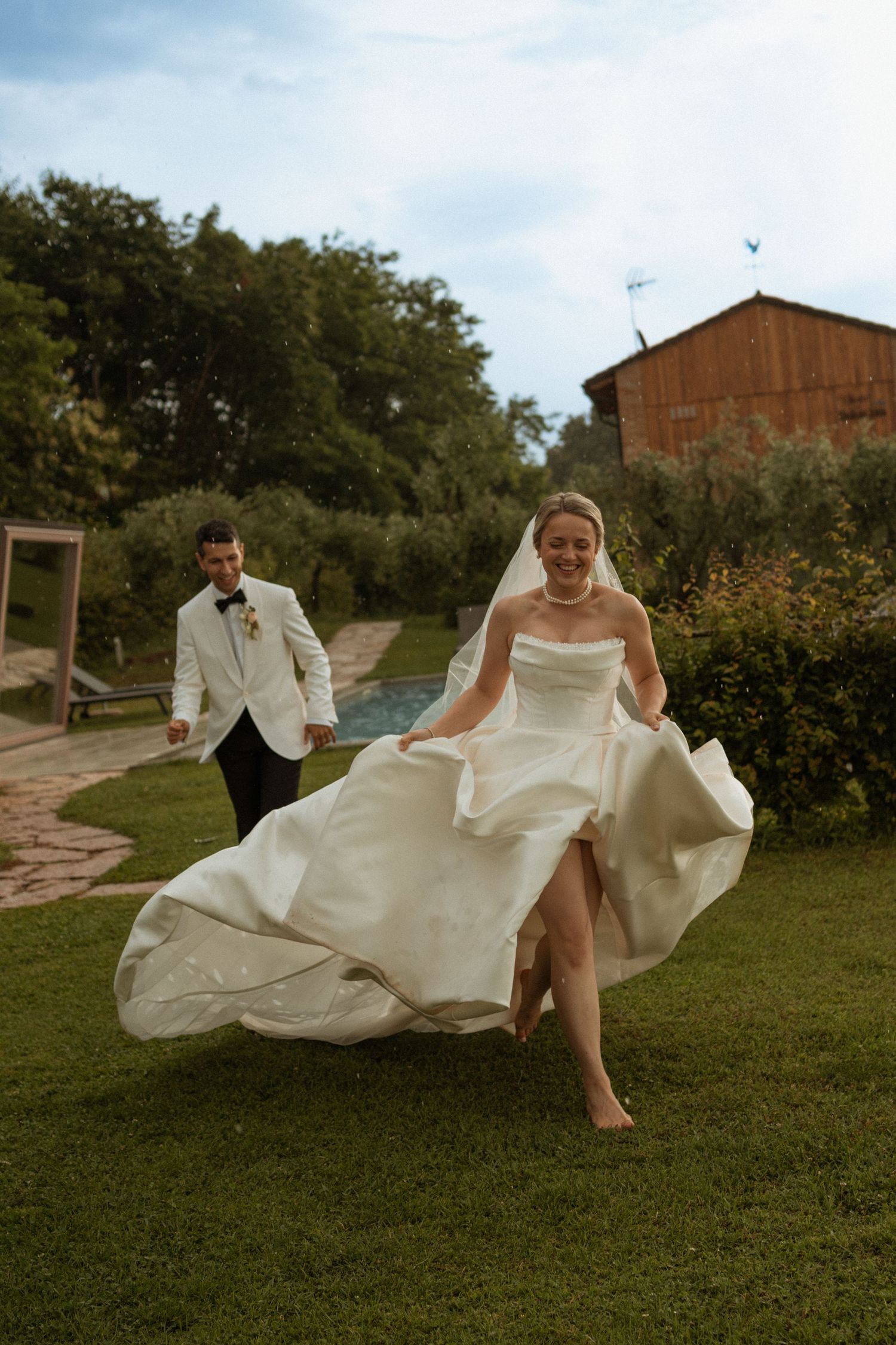 Laughing bride running barefoot across green lawn in flowing white gown as groom follows.