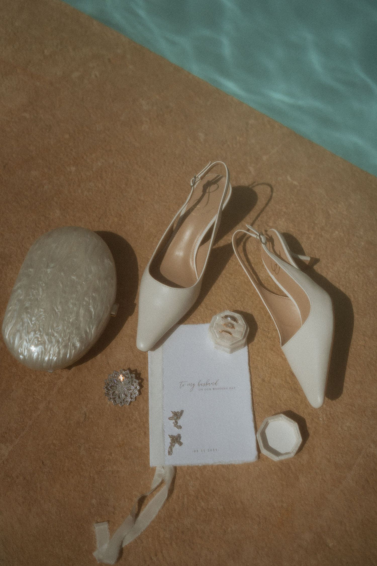 Bridal accessories flatlay featuring white slingback heels, clutch, earrings, and stationery poolside.