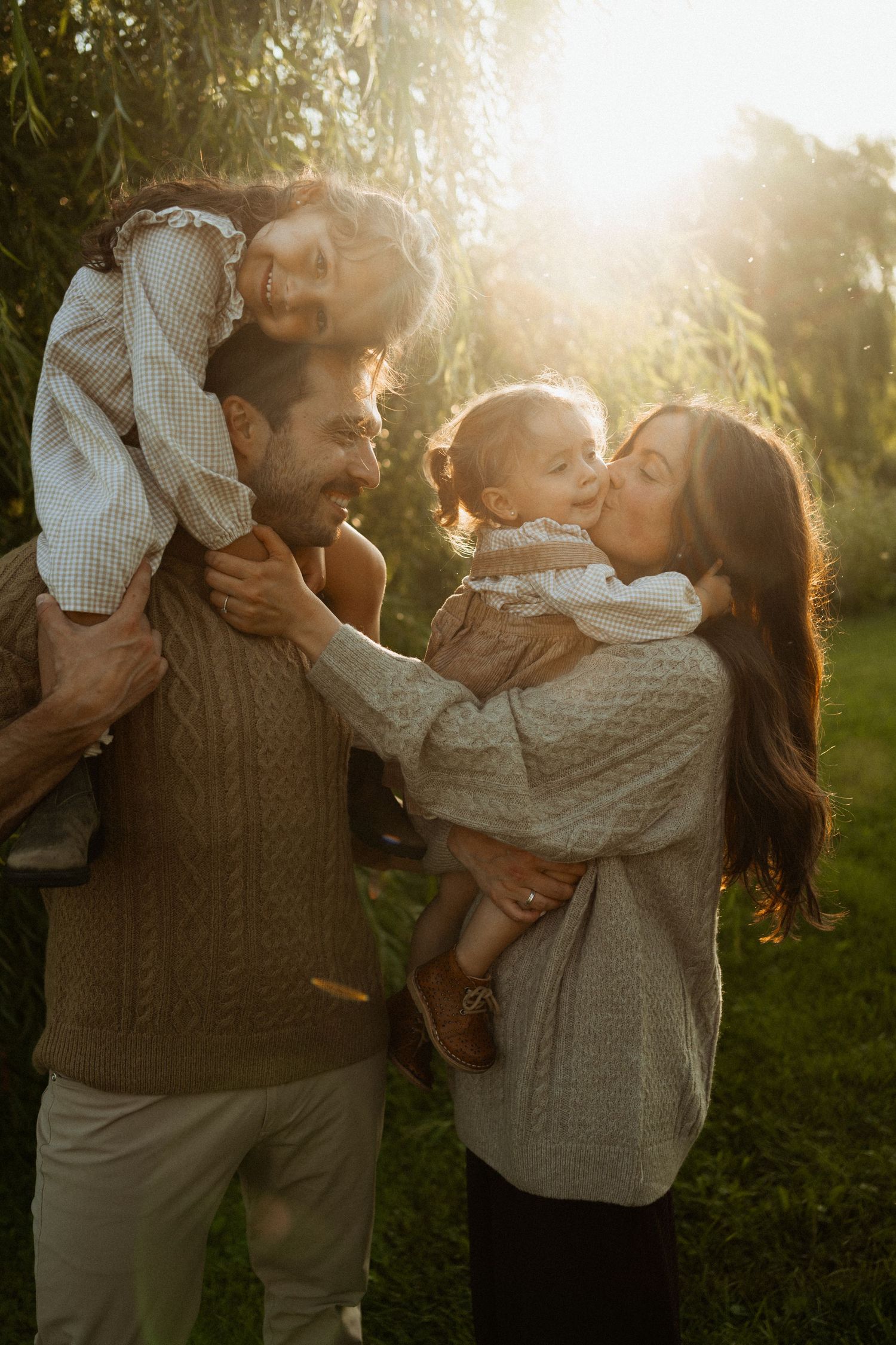 A joyful family of four enjoys a golden hour outdoor moment, with dad carrying a child on his shoulders while mom kisses a toddler.