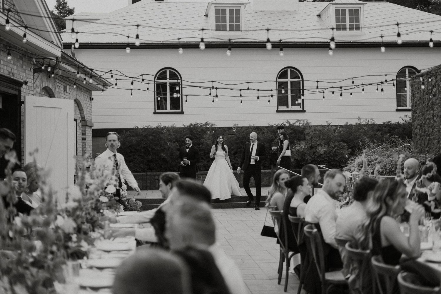 Black and white outdoor wedding reception with string lights, bride and groom standing before seated guests at elegant venue.