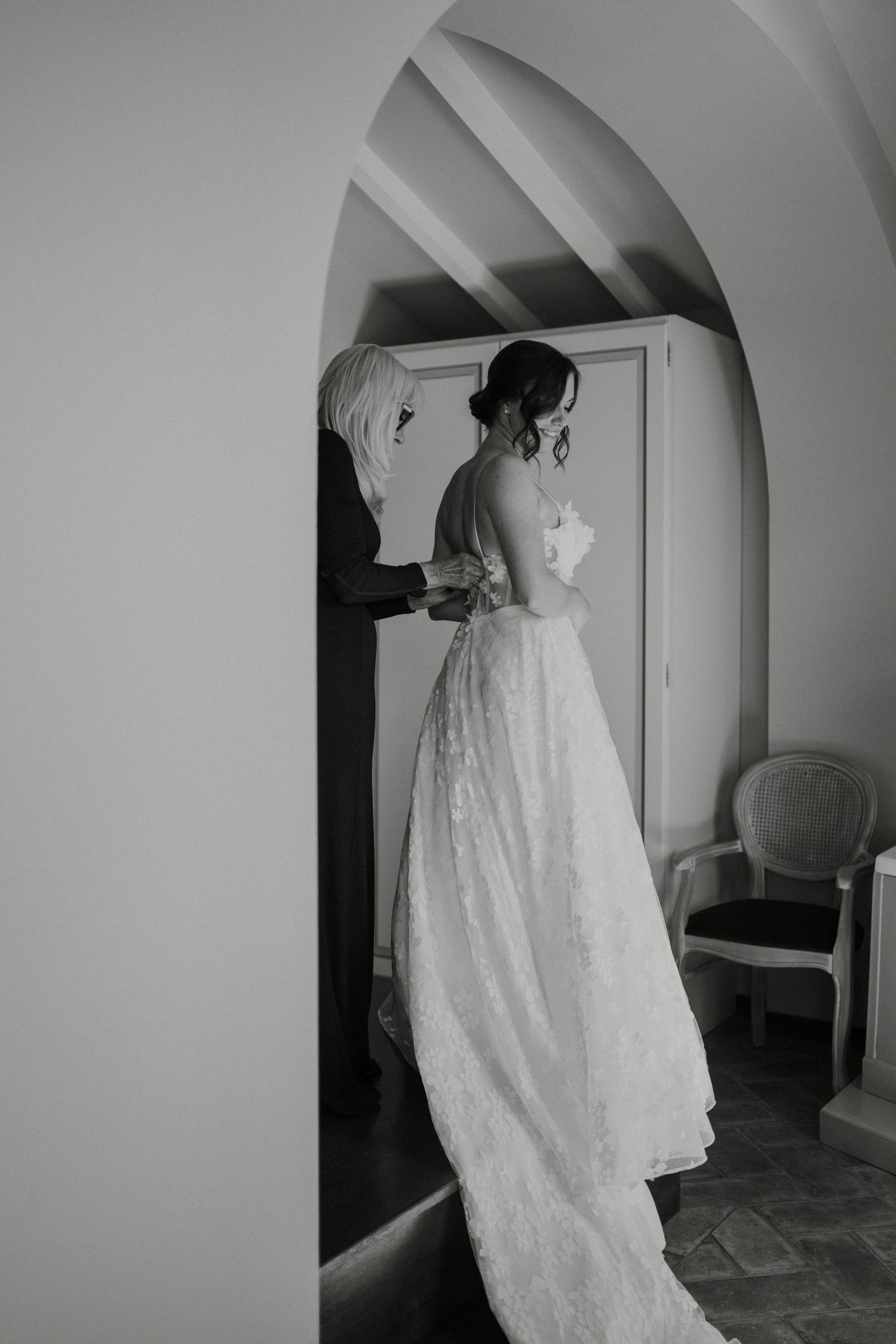 Black and white photo of a bride in lace gown being helped with her dress by a woman in an arched doorway.