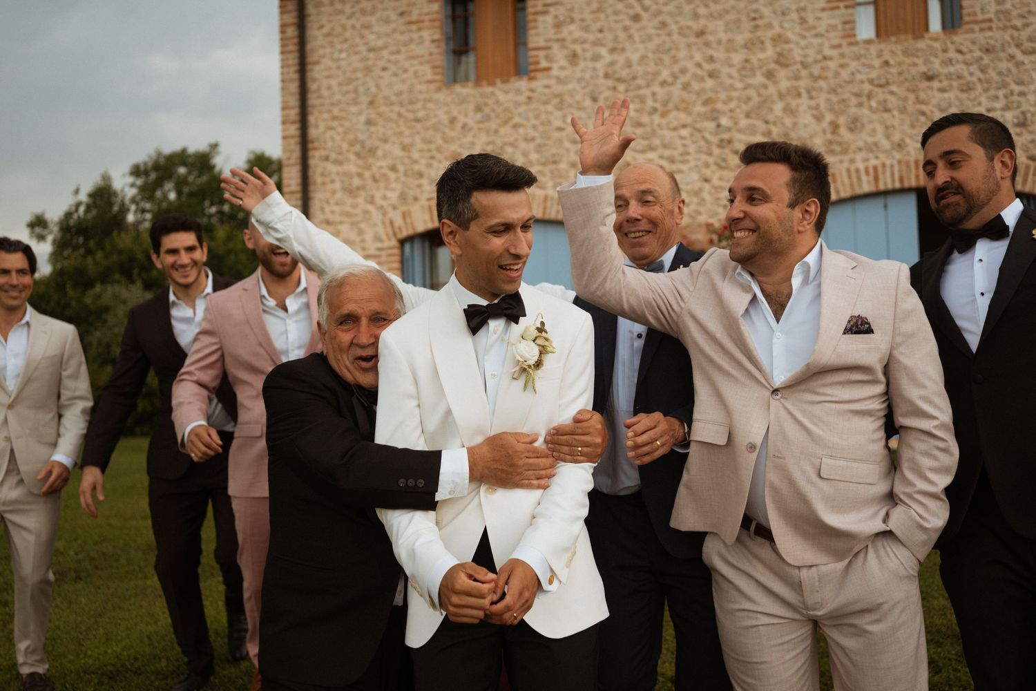 Groom in white tuxedo celebrating with groomsmen outside a rustic stone venue.