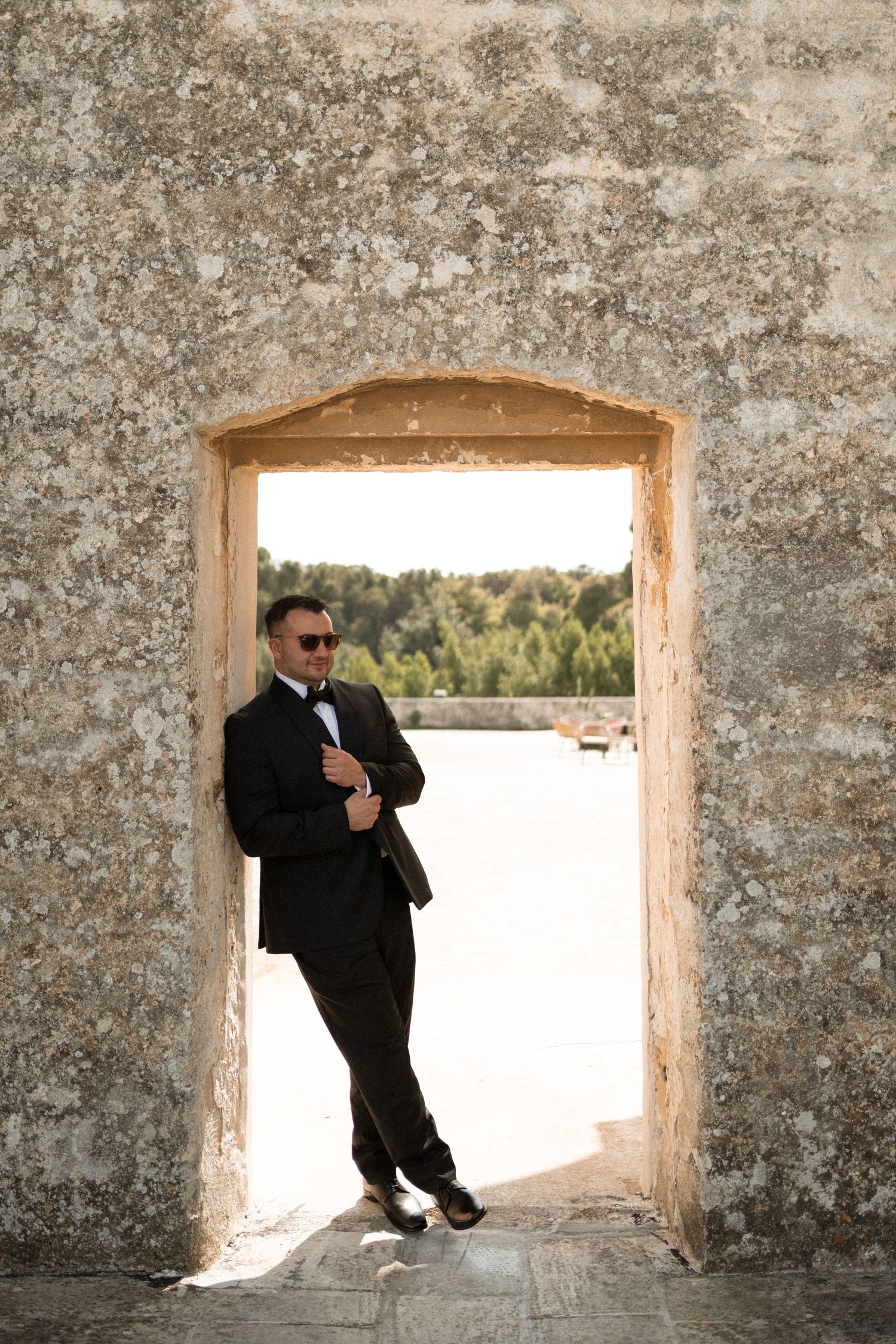 Elegant groom in black tuxedo and sunglasses posing in a rustic stone archway outdoors.