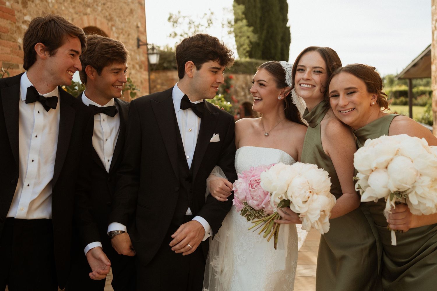 Bride and groom smile with wedding party in formal attire, bridesmaids holding white bouquets at outdoor venue.