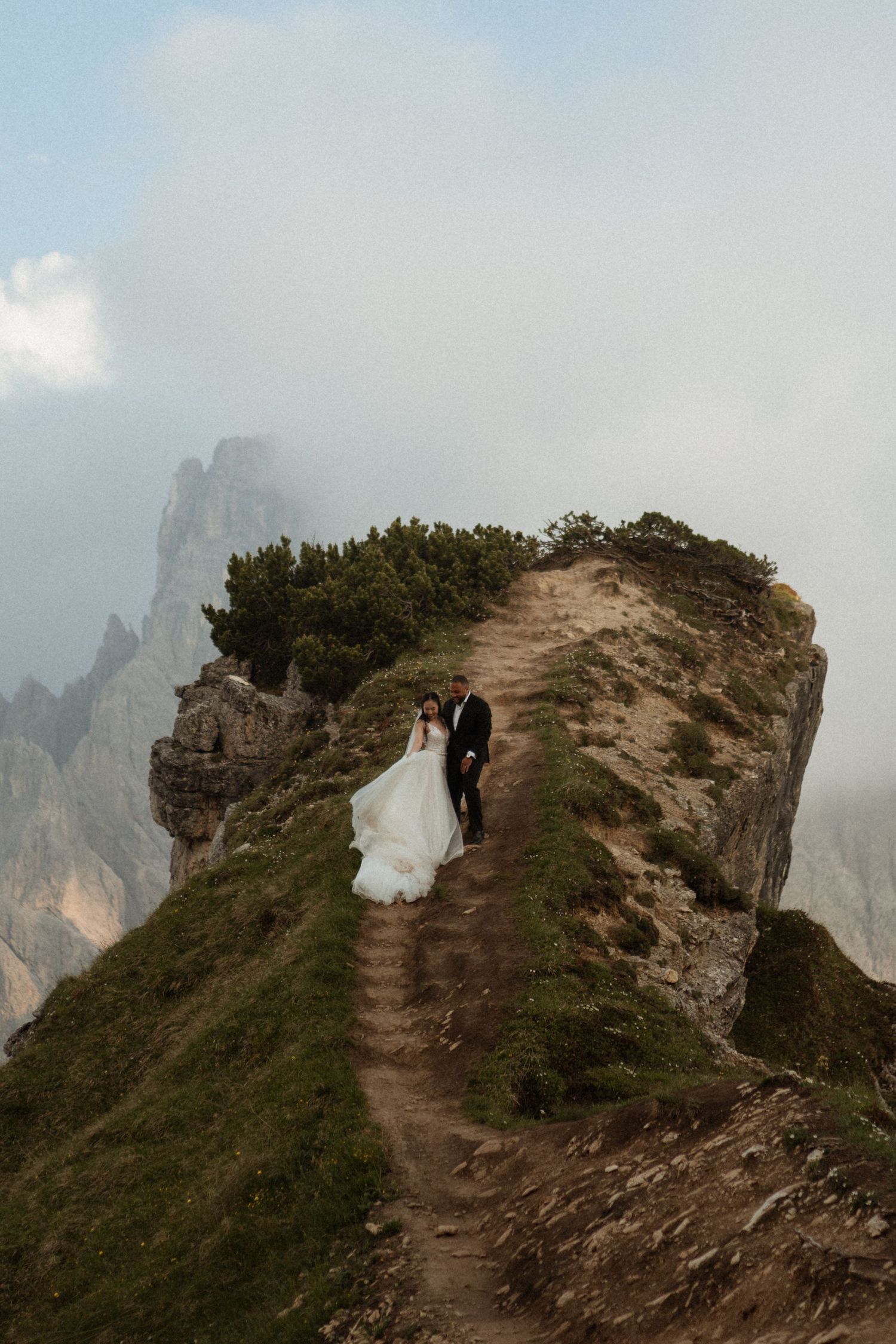 Bride and groom stand on a misty mountain cliff edge in the Dolomites, wedding dress flowing in the wind.