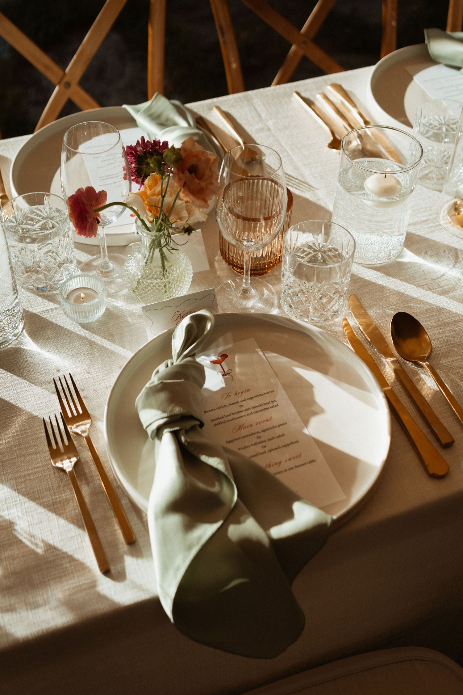 Elegant wedding table setting with gold cutlery, sage green napkin, floral centerpiece, and glassware on a white linen tablecloth.