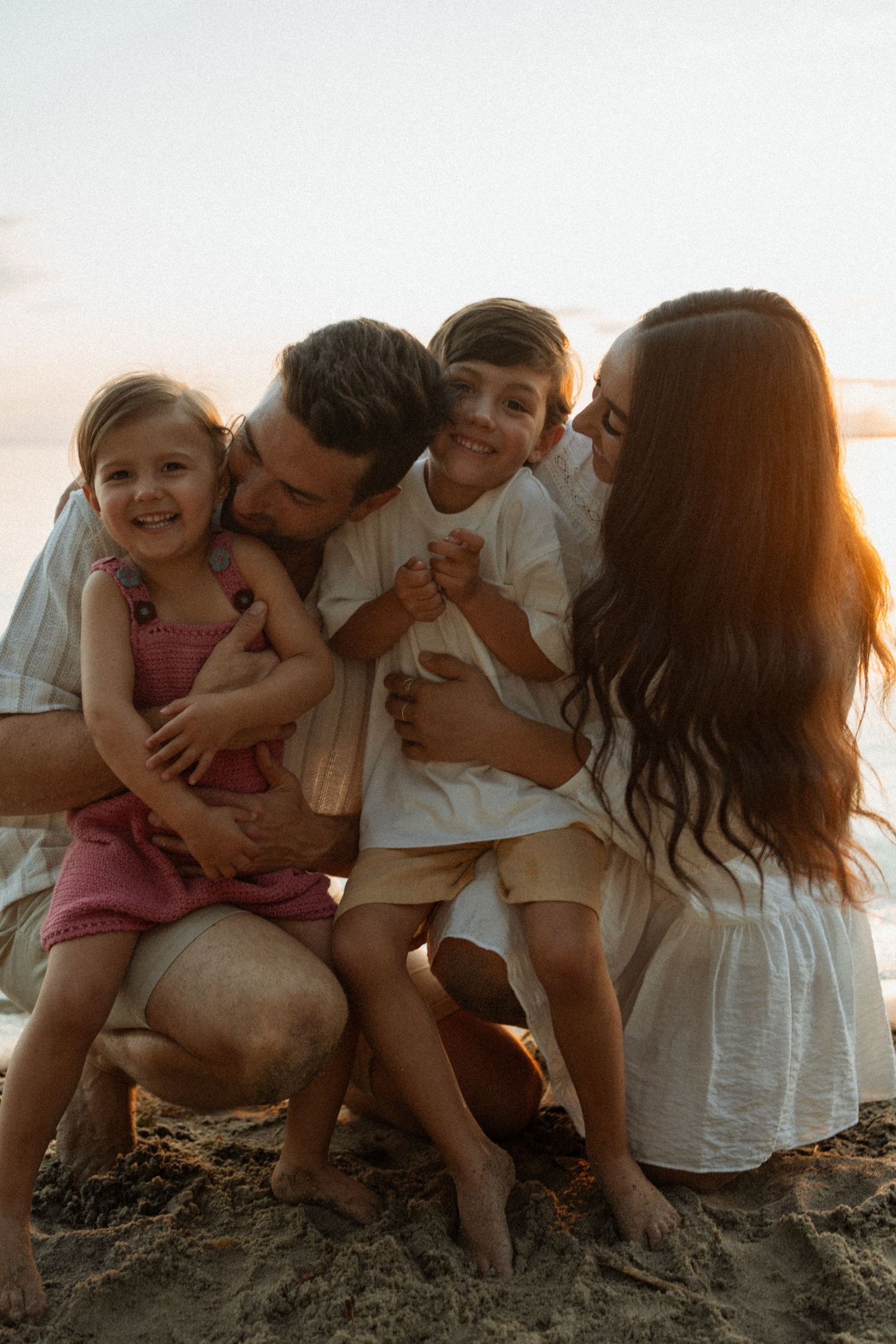 Happy family of four enjoying a golden sunset beach moment, parents hugging smiling children on sandy shore.