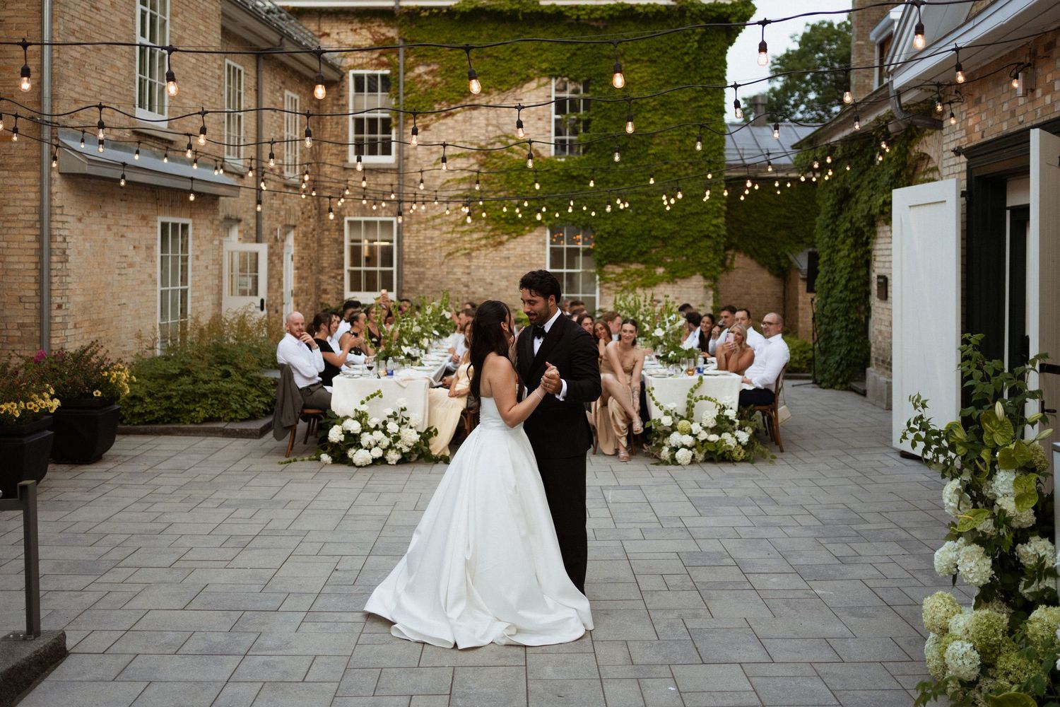 Couple shares first dance in elegant outdoor courtyard wedding with string lights, floral arrangements, and ivy-covered brick venue.