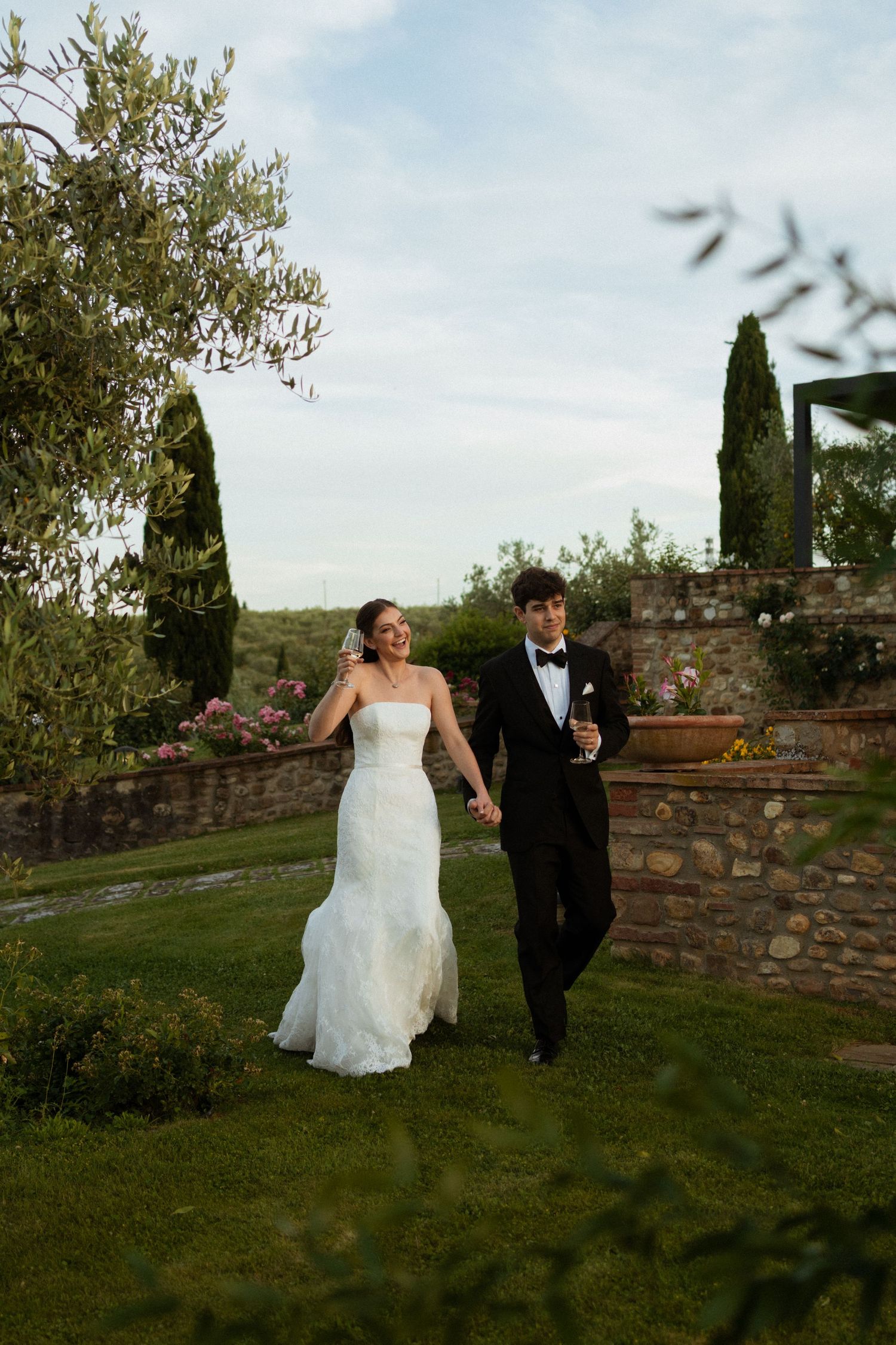 Bride and groom walking hand in hand through lush Tuscan garden with cypress trees.