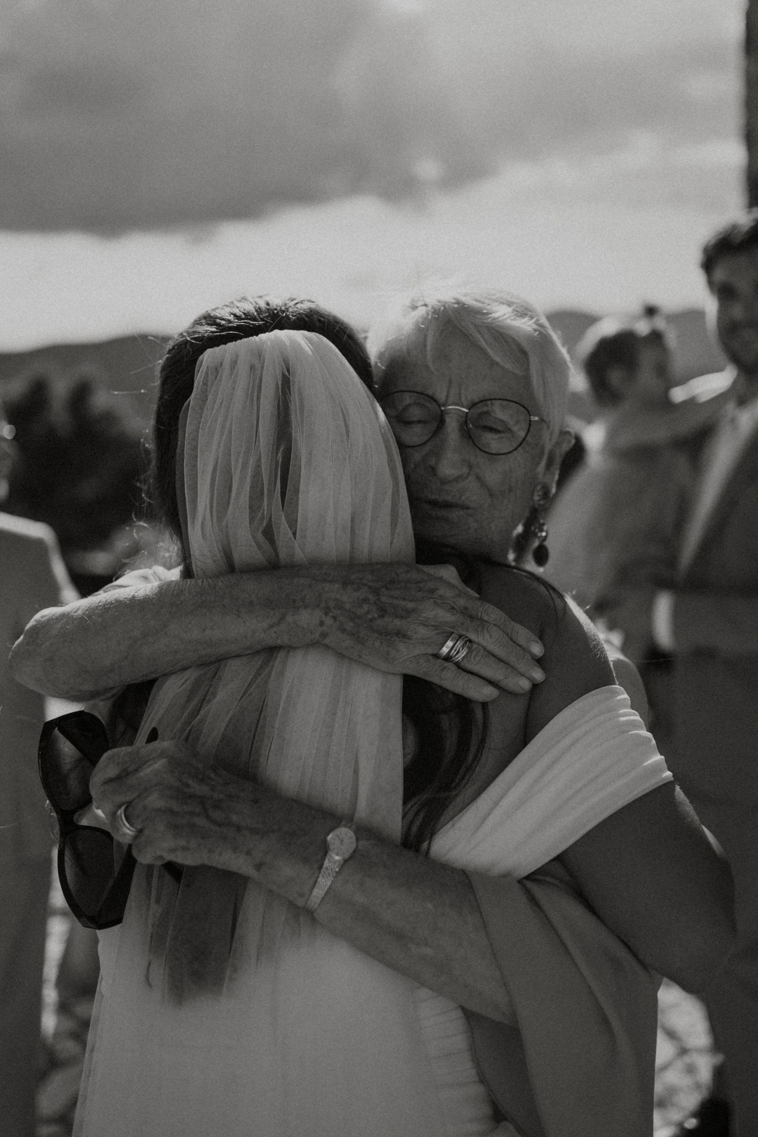 Emotional black and white photo of bride in veil receiving warm hug from elderly wedding guest.