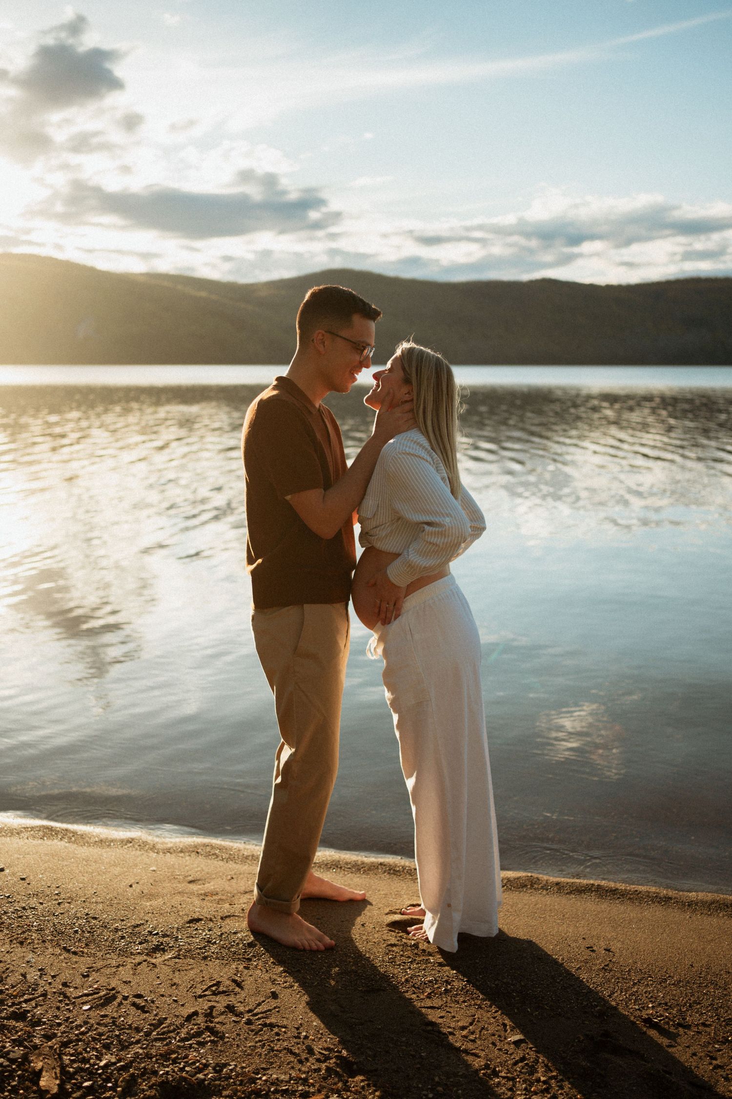 Couple sharing a romantic moment on a sandy lakeside beach at golden sunset with mountain backdrop.