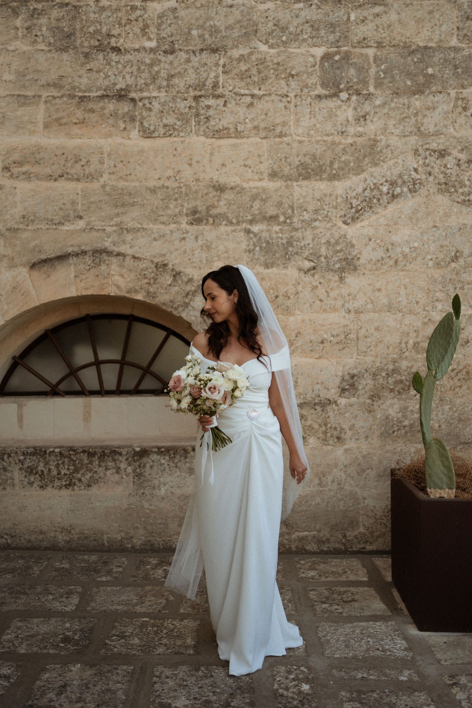 Bride in white off-shoulder gown and veil holds bouquet beside stone wall with cactus.