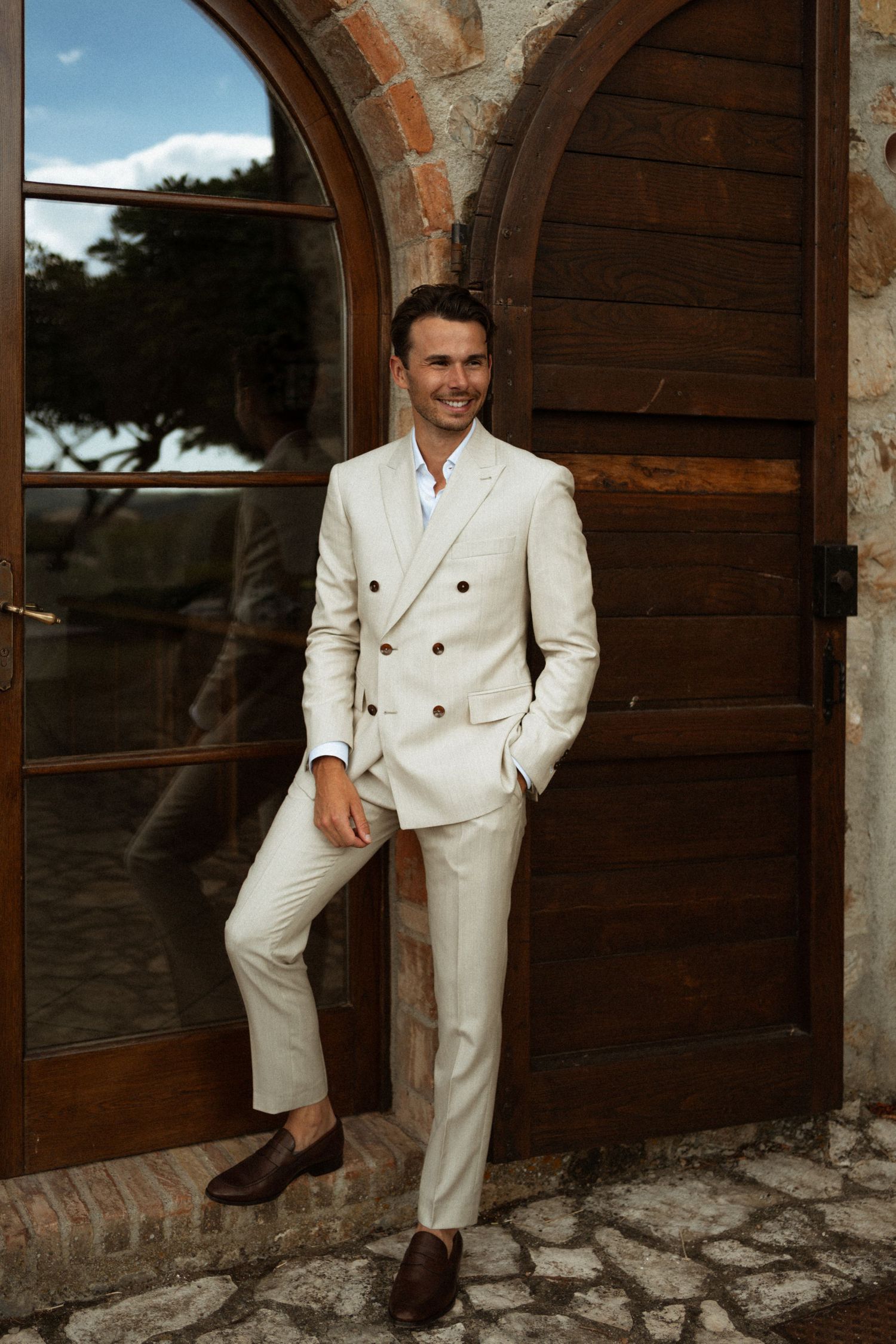 Smiling groom in cream double-breasted suit leaning against rustic wooden arched door at Tuscan villa.