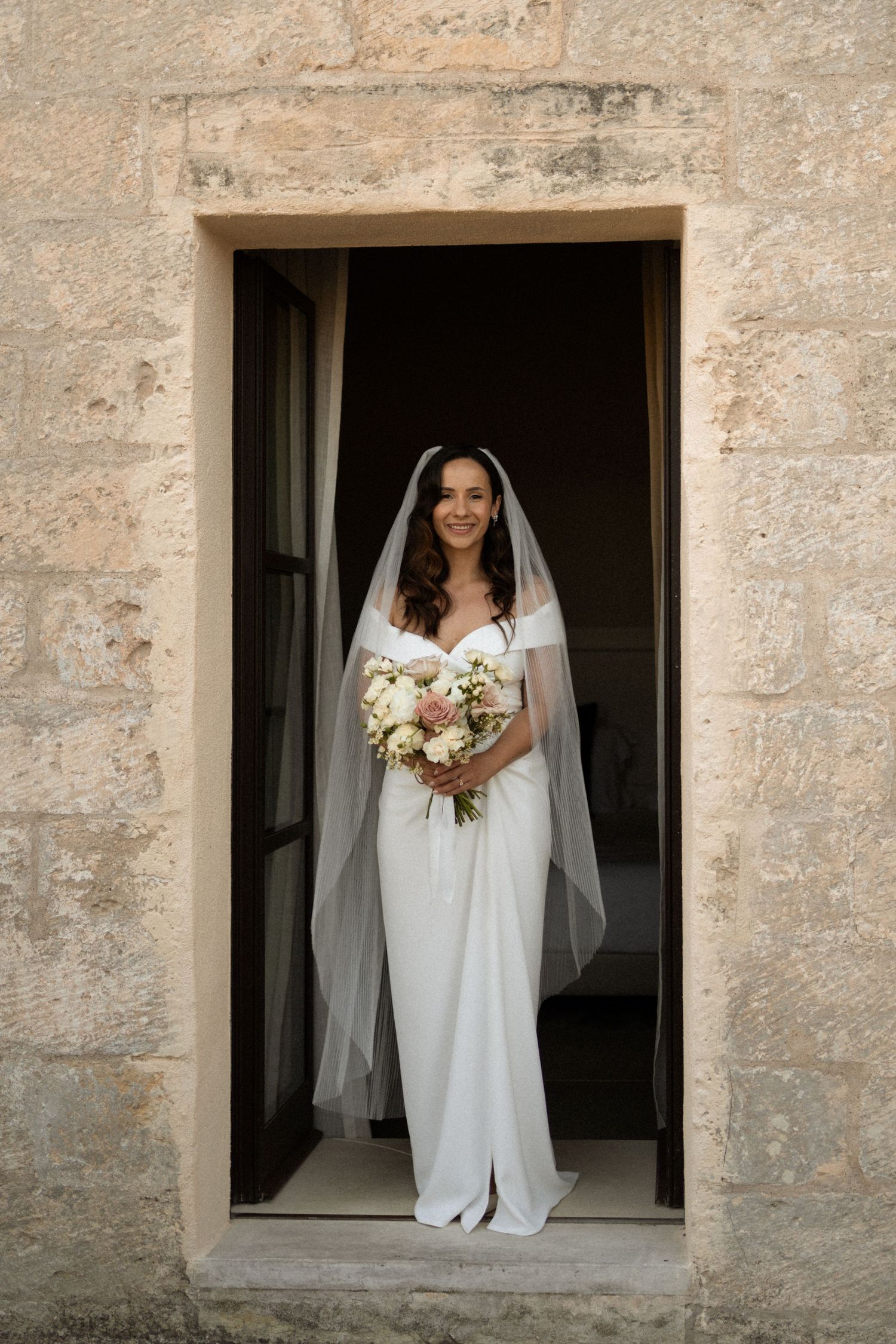 Beautiful bride in white gown and veil holding bouquet standing in a stone doorway.