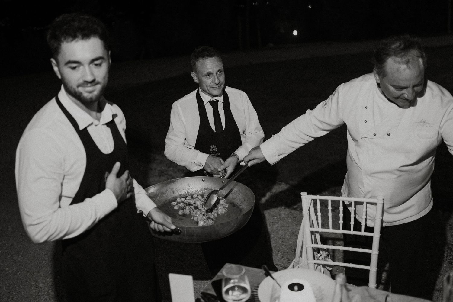 Black and white photo of three men preparing food outdoors at night, with a large wok pan at an event.