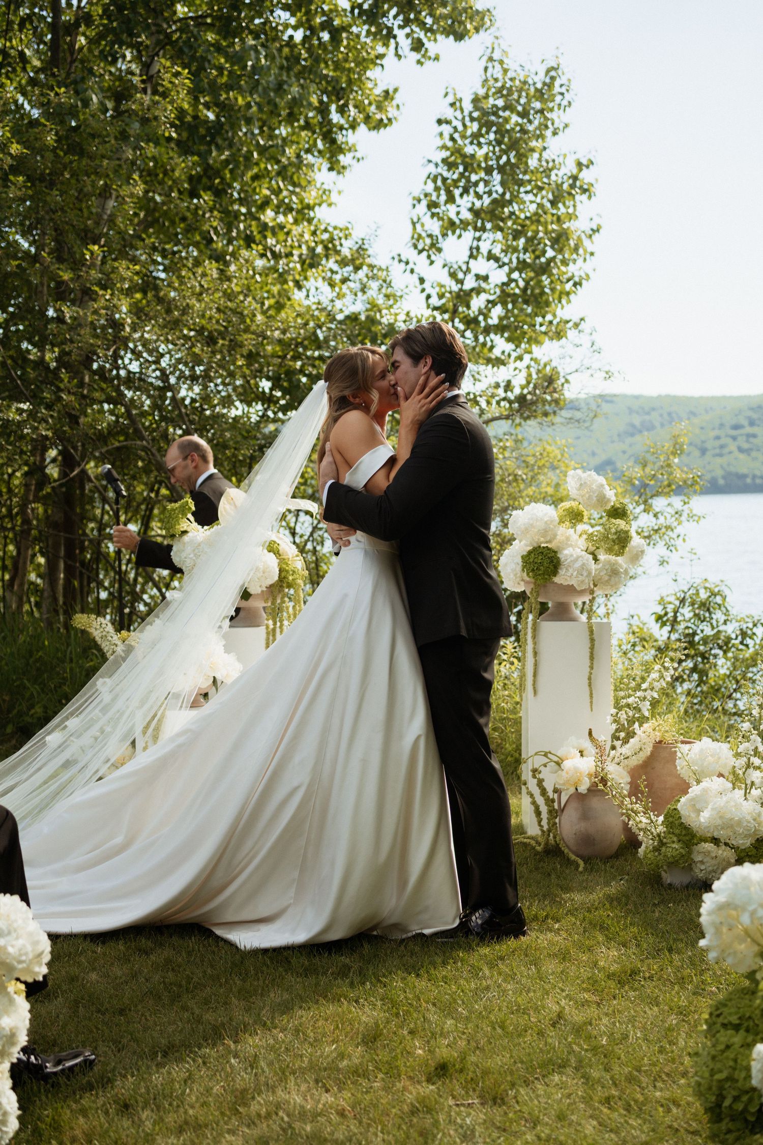 Bride and groom share first kiss at outdoor lakeside wedding ceremony surrounded by white floral arrangements.