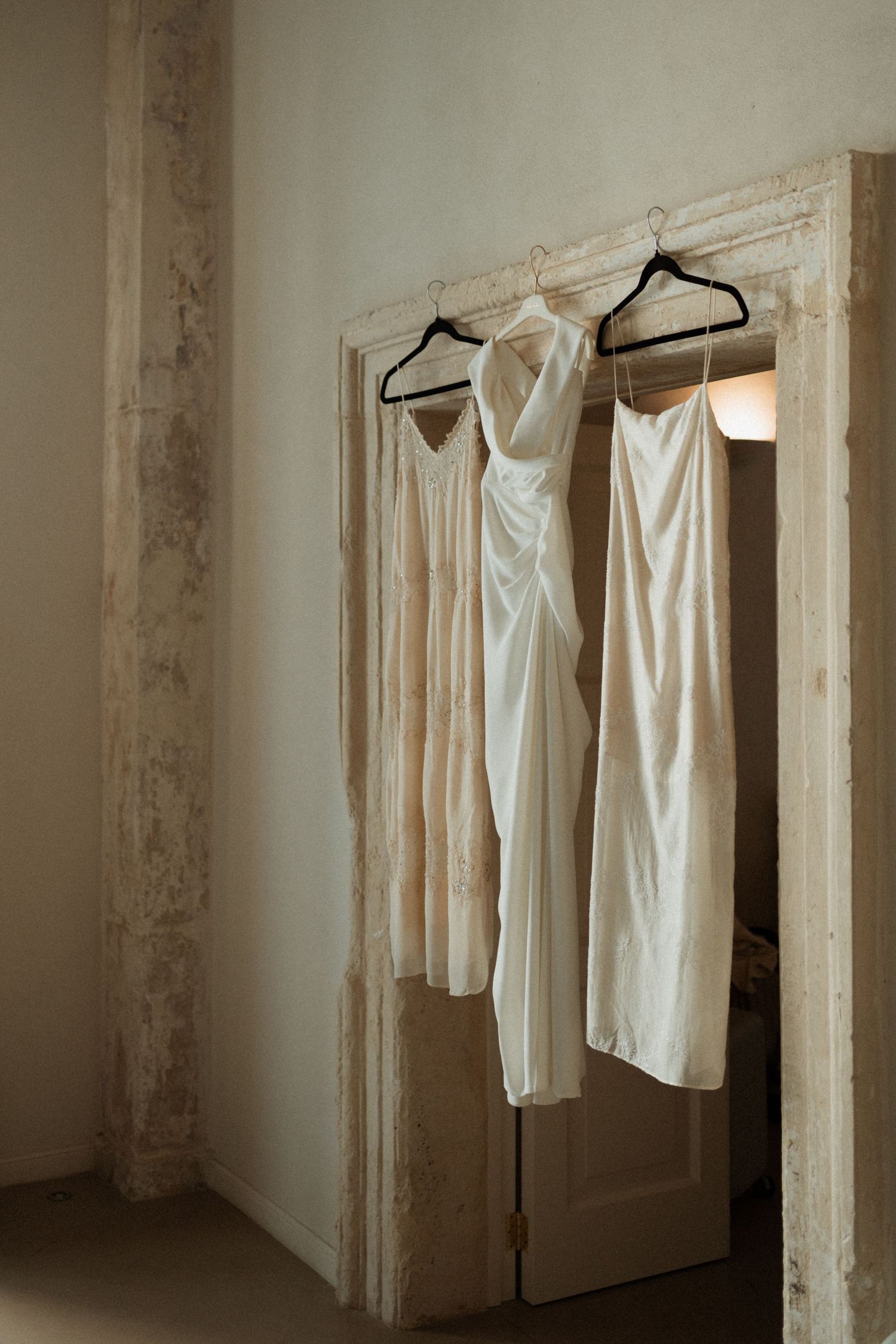 Two white wedding dresses hanging on black hangers in a rustic stone doorway.