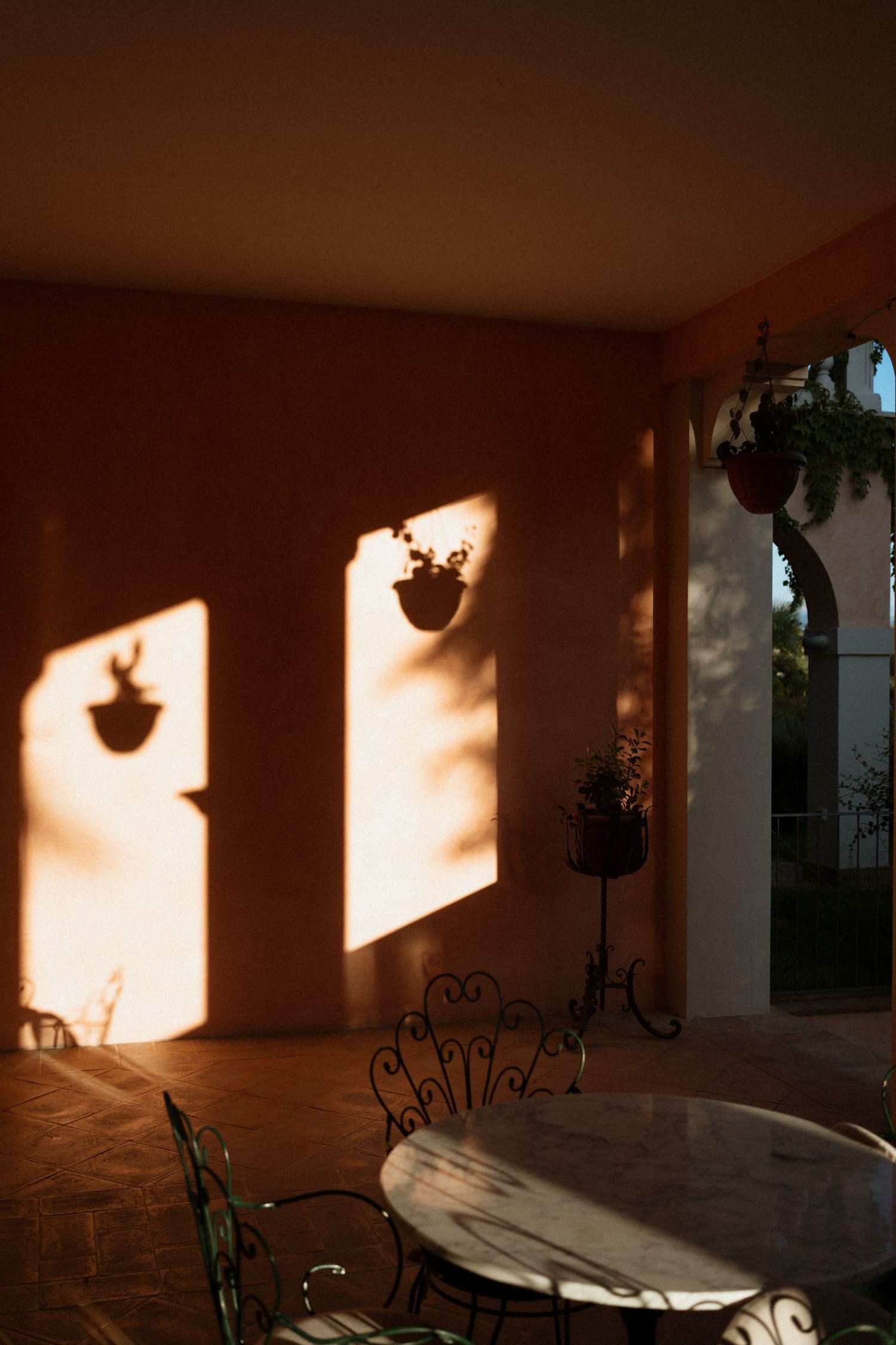 Warm sunset light casts dramatic shadows of hanging plants on an orange terrace wall with wrought iron furniture.