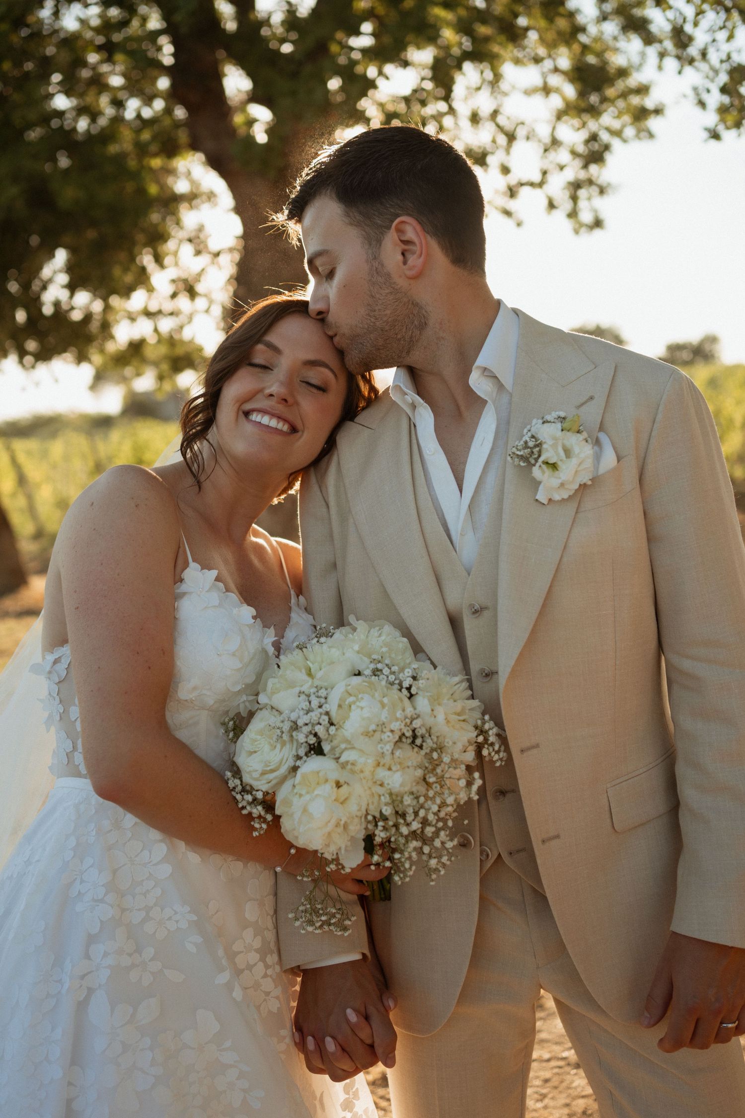 A smiling bride holding white bouquet leans on groom in beige suit kissing her forehead at golden hour.