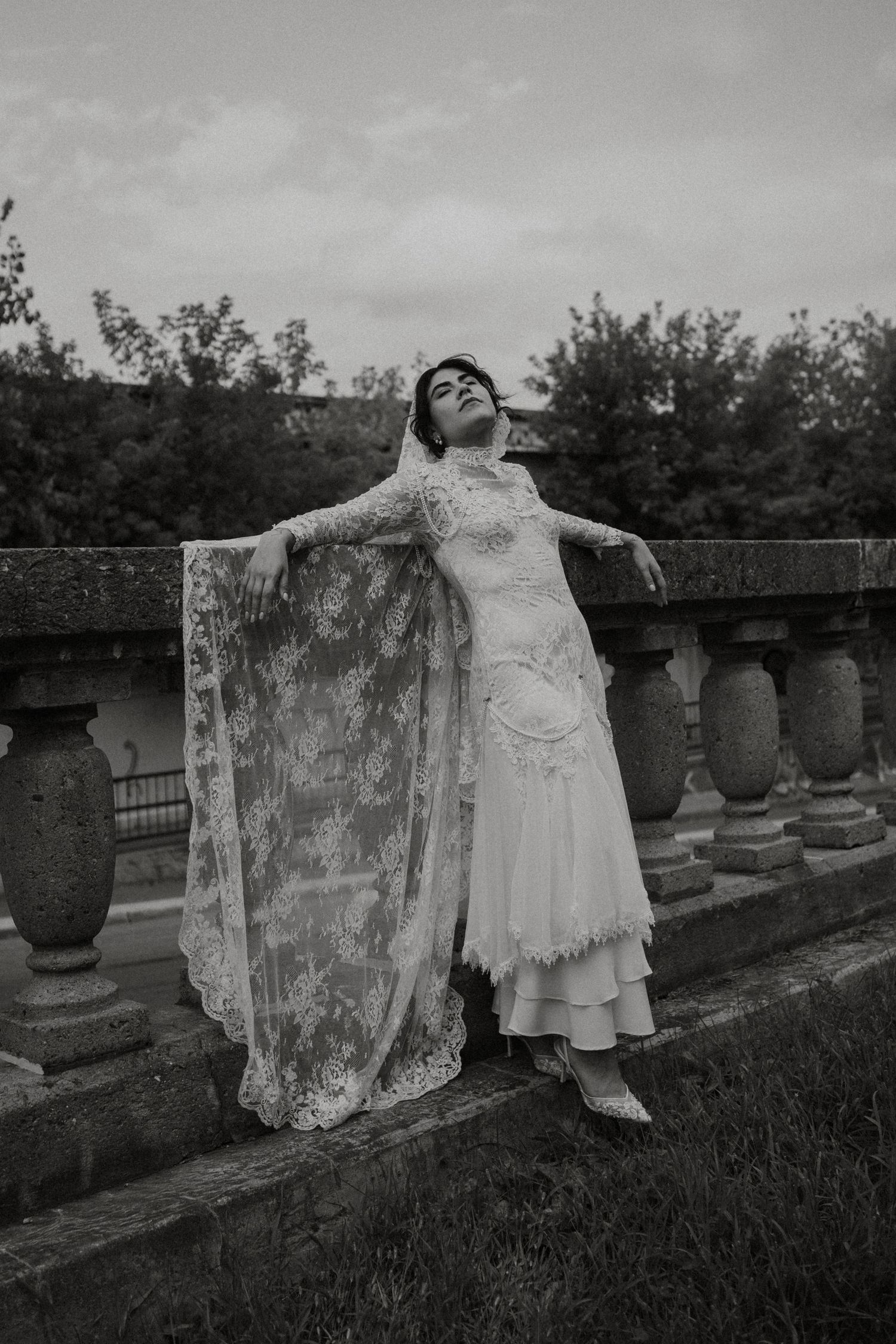 Black and white photo of a bride in lace gown and veil posing gracefully against a stone balustrade outdoors.