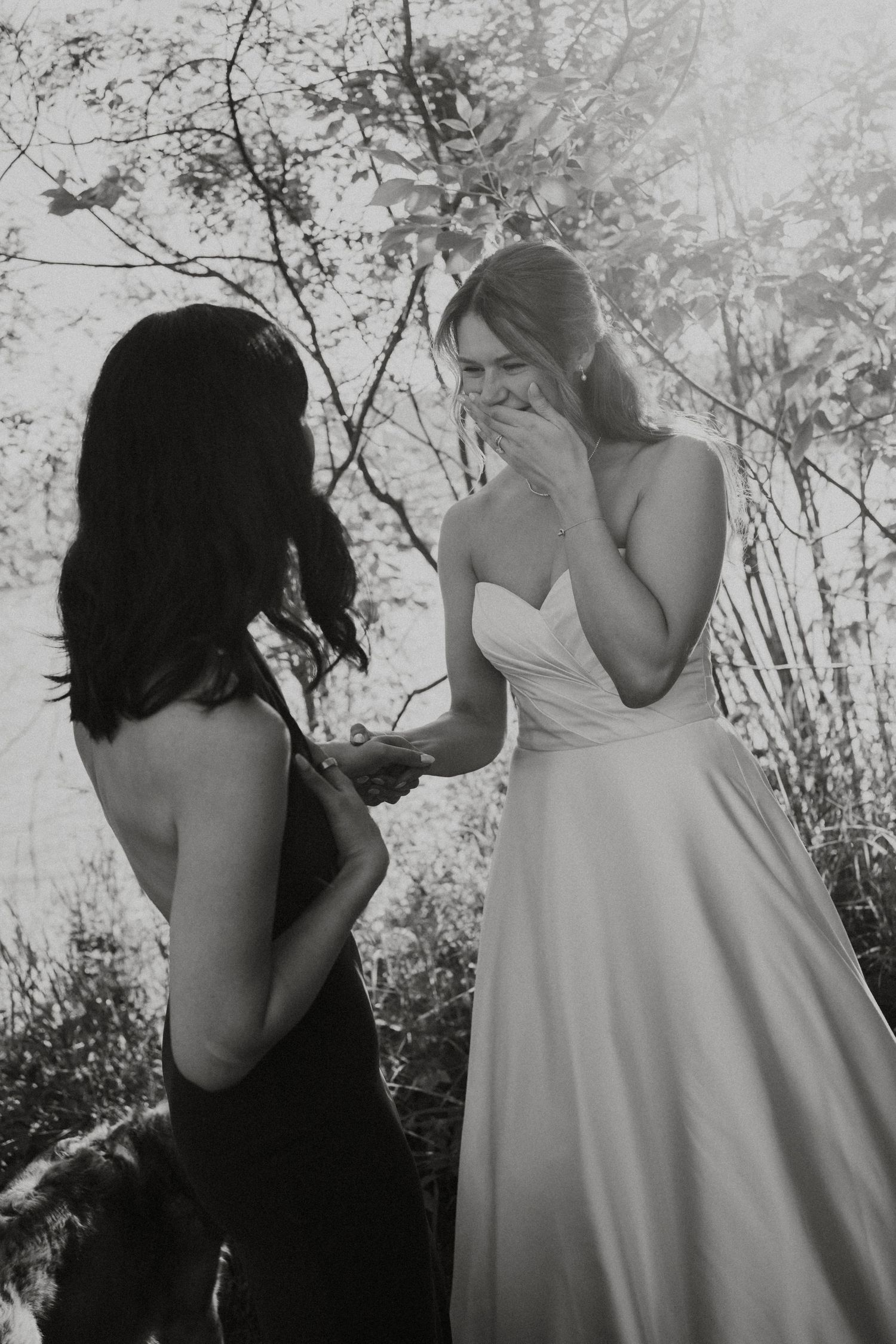 Black and white photo of emotional bride covering mouth while holding hands with woman in outdoor setting.