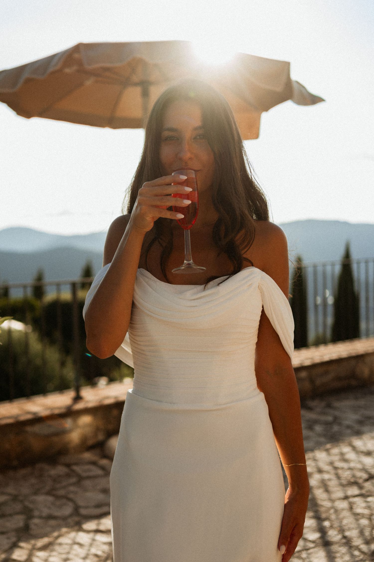Bride in white off-shoulder gown sipping wine at sunset with mountain landscape in background.
