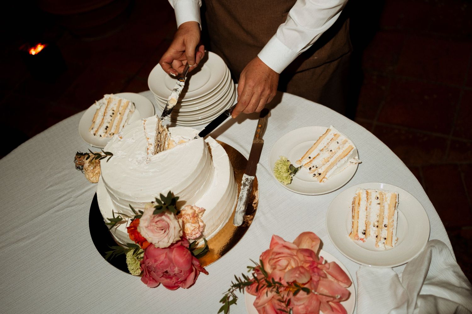Elegant wedding cake being cut with floral decorations and plated slices on a white tablecloth.
