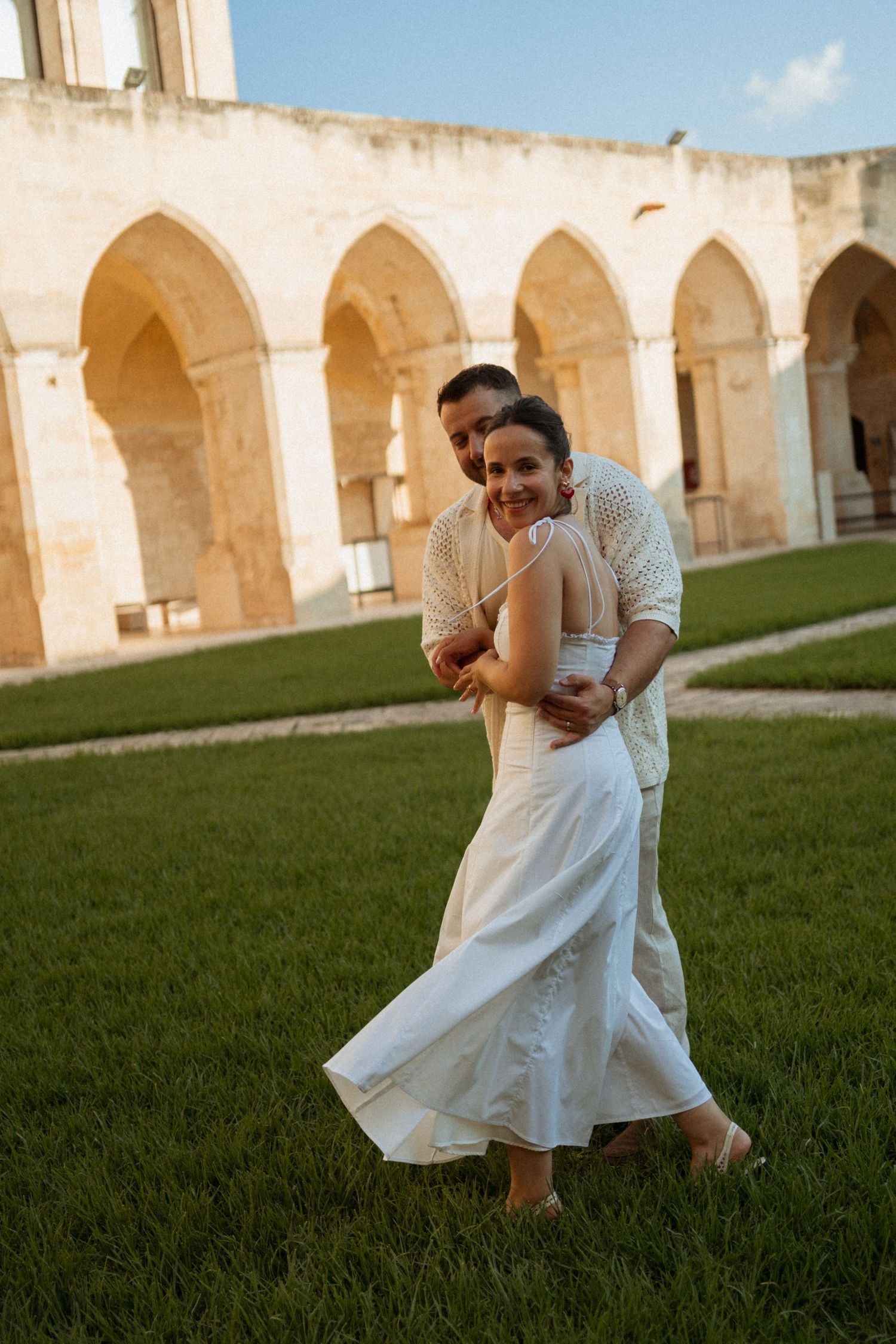 Couple dancing on green grass in front of ancient stone arches at golden hour, woman in white dress.