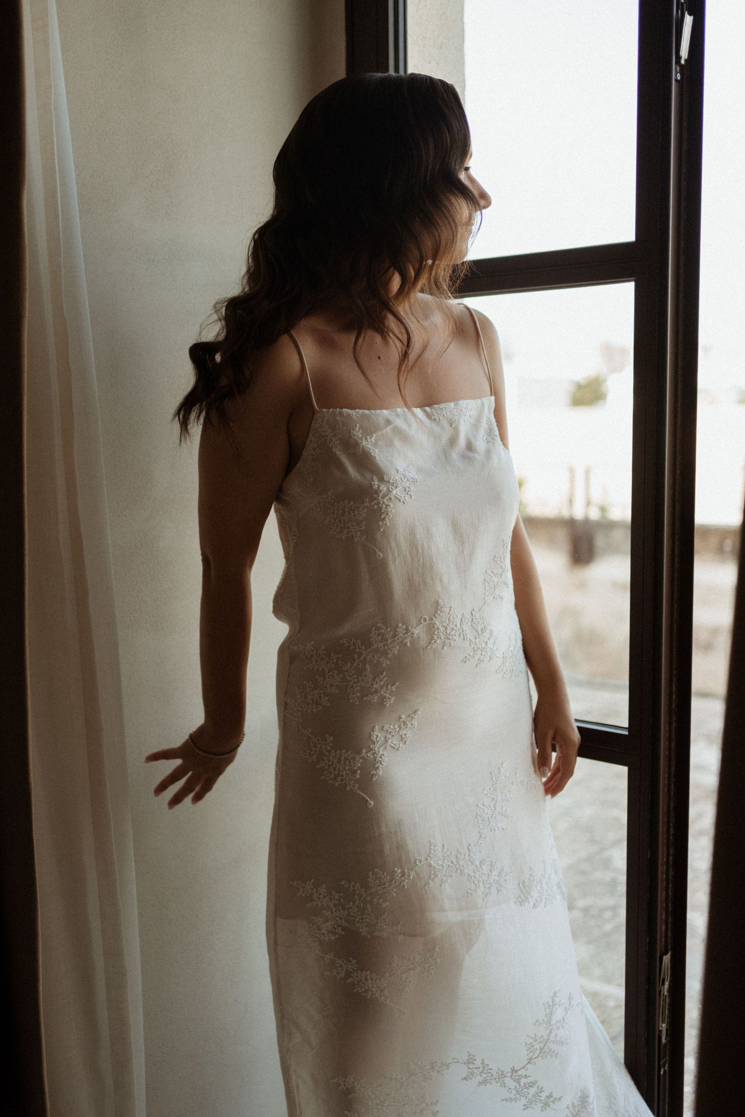Bride in elegant white lace wedding dress standing at an open window in soft light.