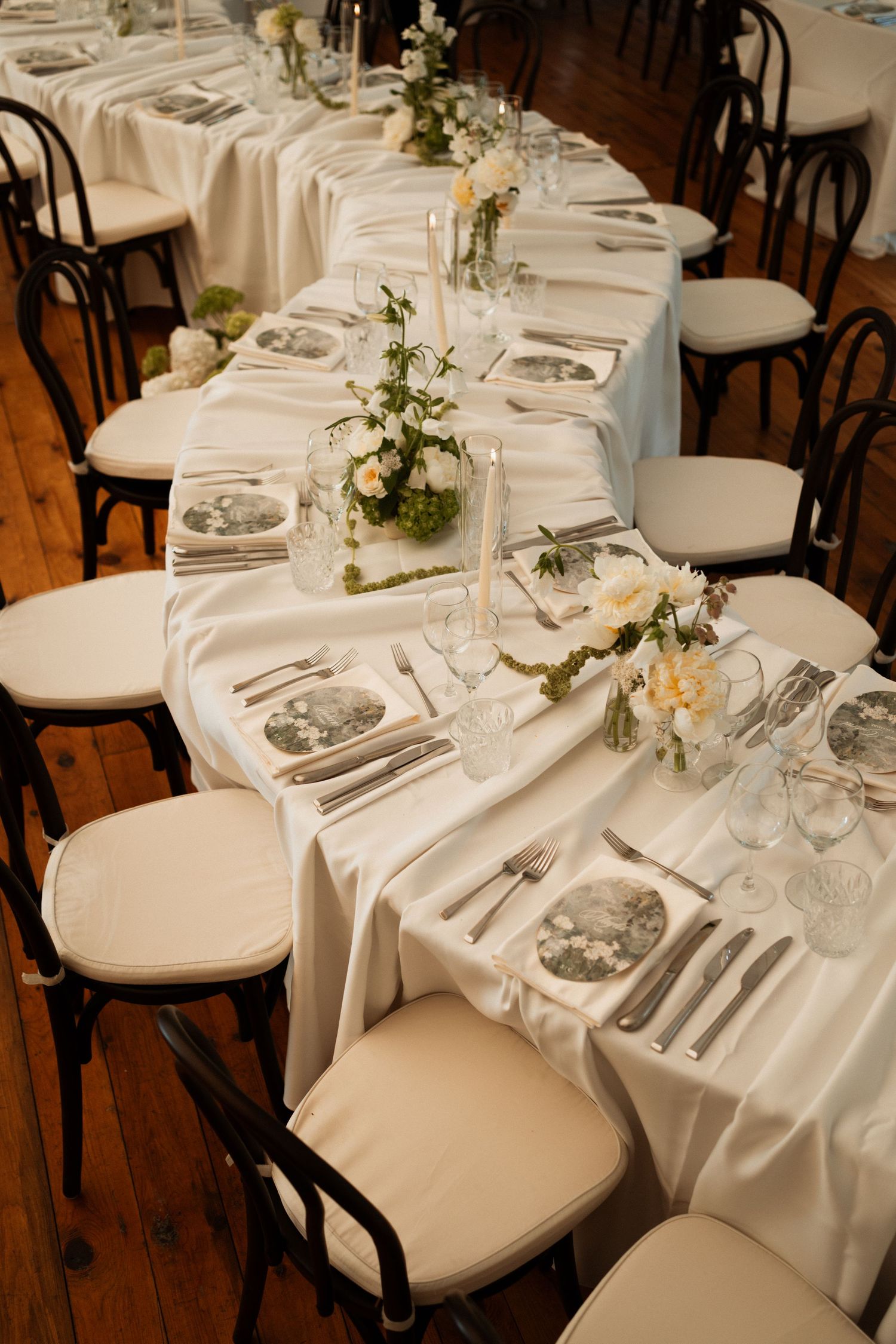Elegant wedding reception table with white satin runner, floral centerpieces, fine china plates, and dark bentwood chairs.