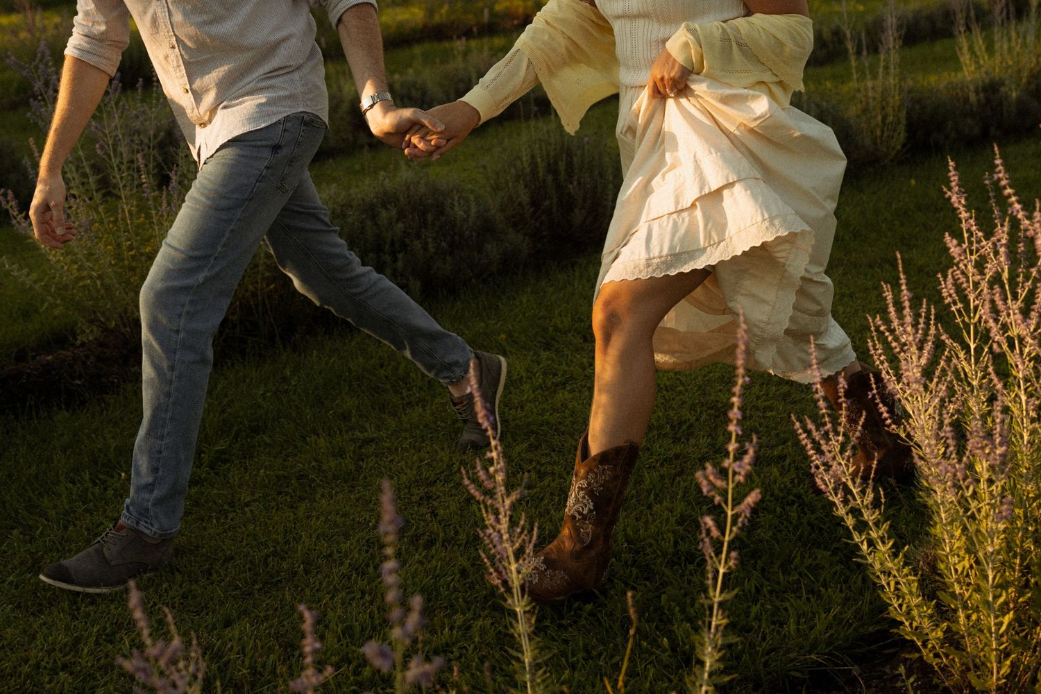 Couple holding hands running through a lush green field with wildflowers at golden hour.