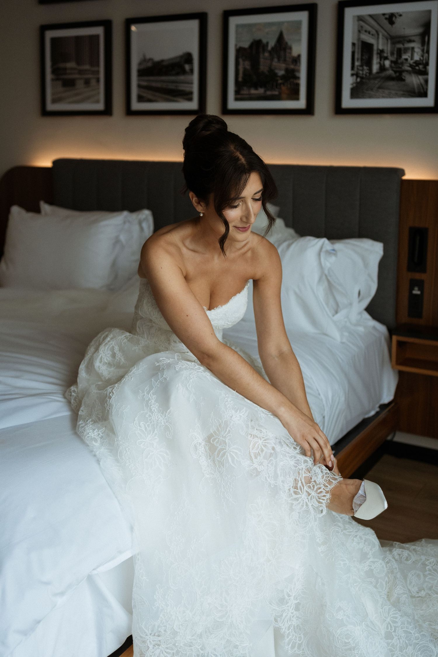 Bride in strapless lace gown putting on wedding shoes while sitting on hotel bed.