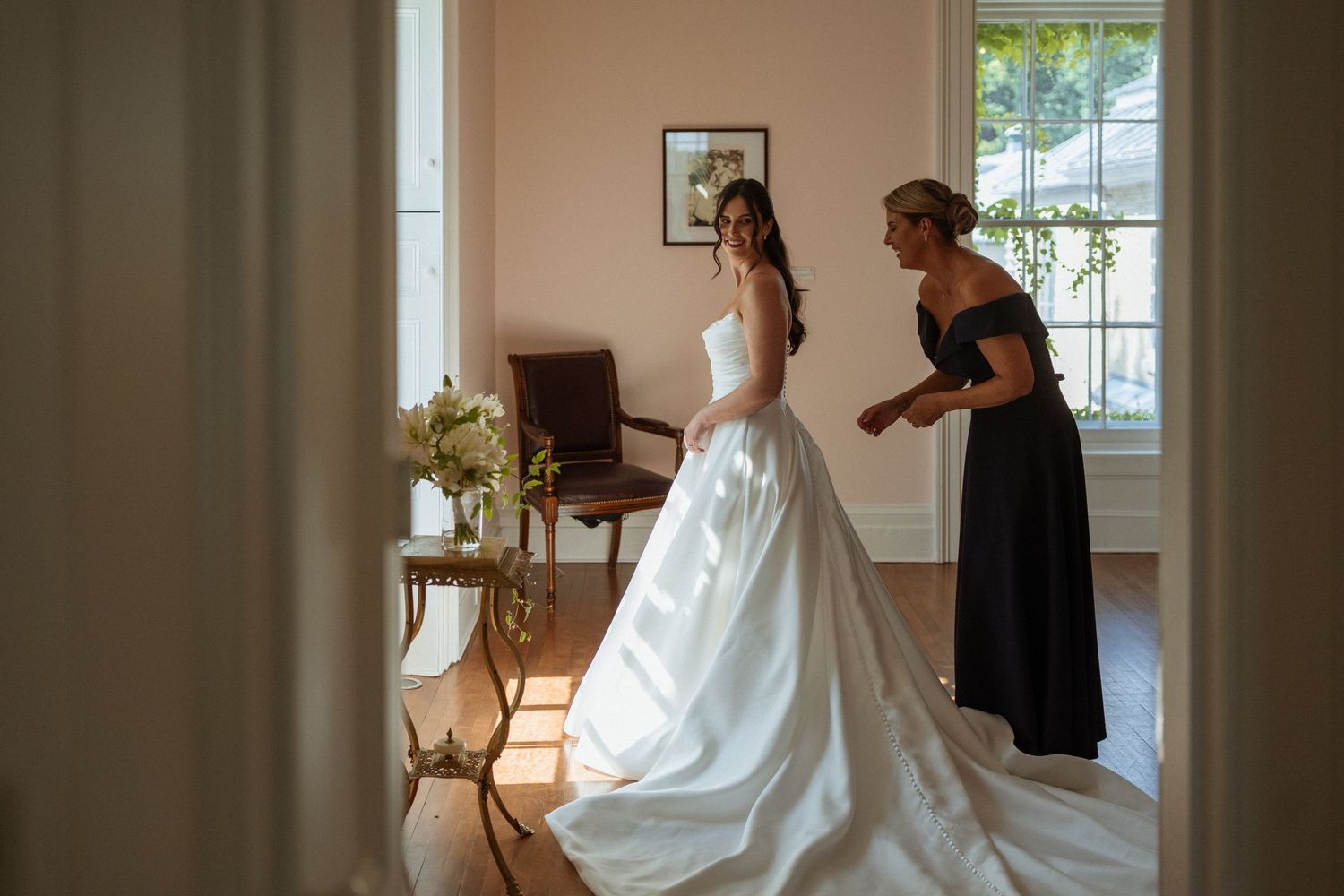 Bride in white ballgown being helped by woman in black dress in elegant sunlit room.