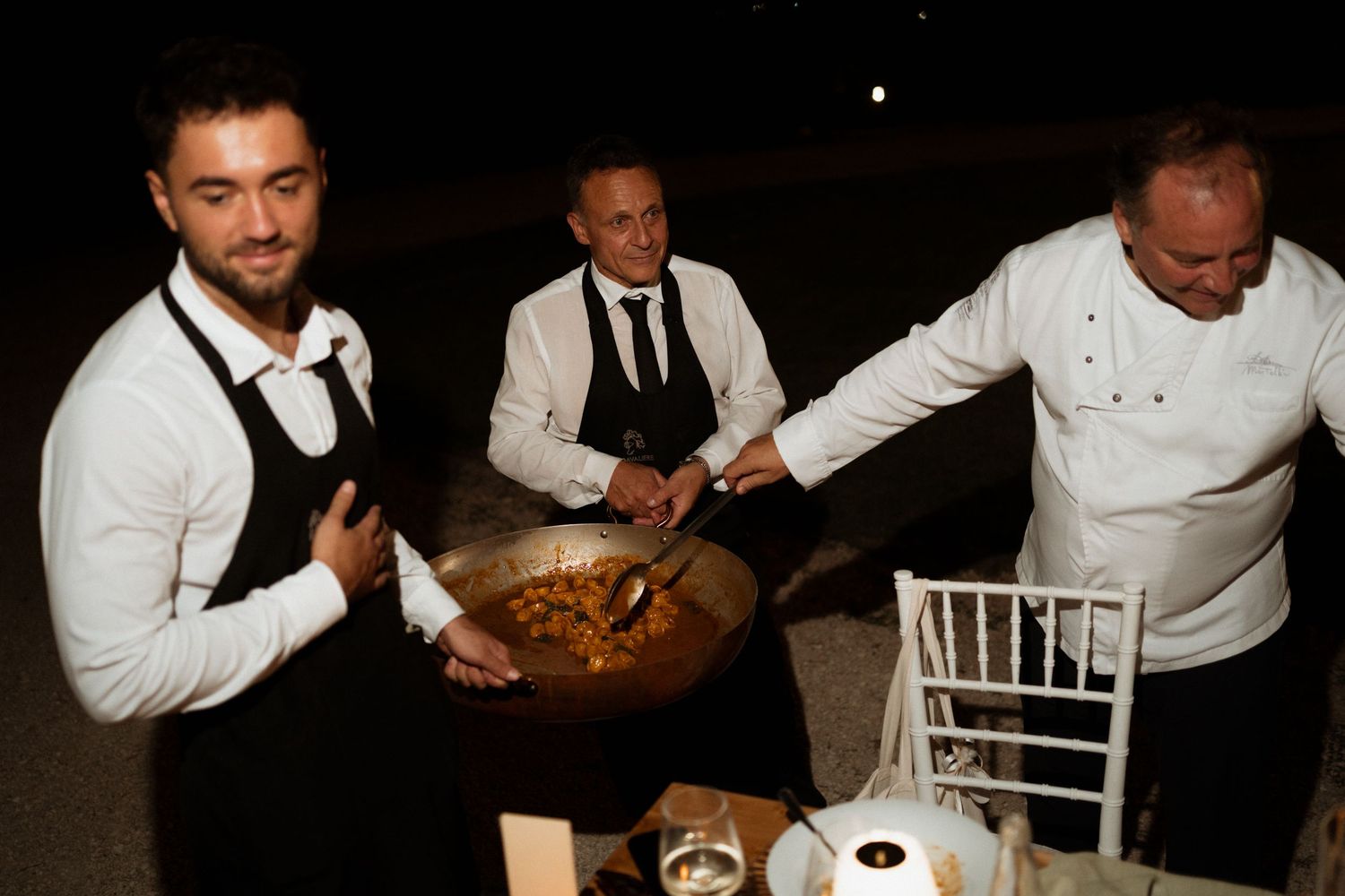 Three catering staff serve food from a large wok at an outdoor evening event, with a chef in white uniform.